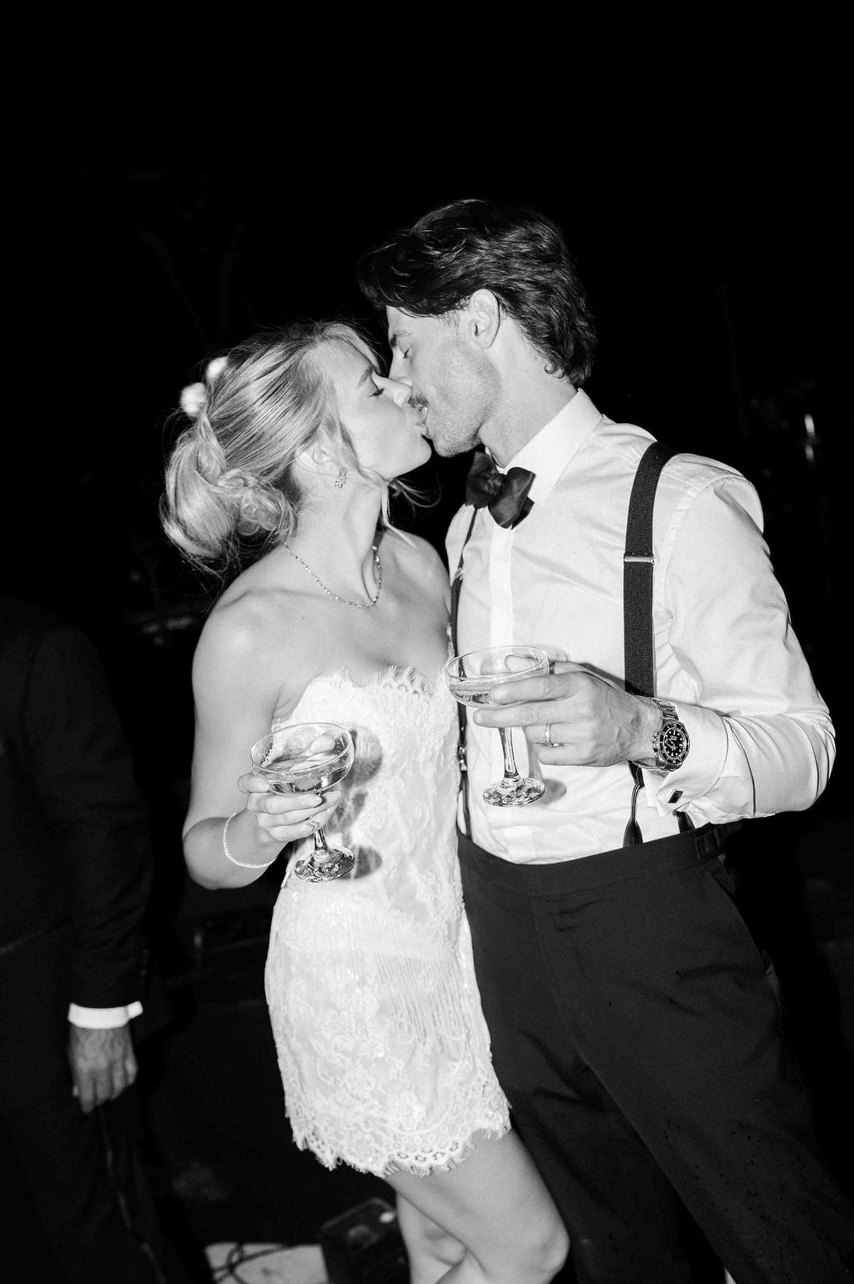 In black and white, the couple kiss while each holding a champagne coupe, the groom wearing braces and a bow tie.
