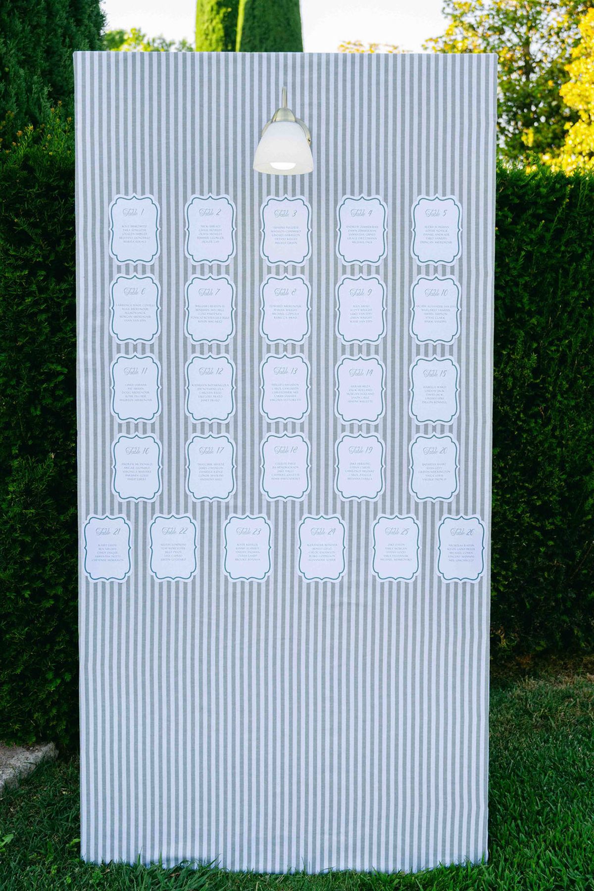 A tall blue and white striped board displays small framed place cards, standing on grass in front of green hedges.