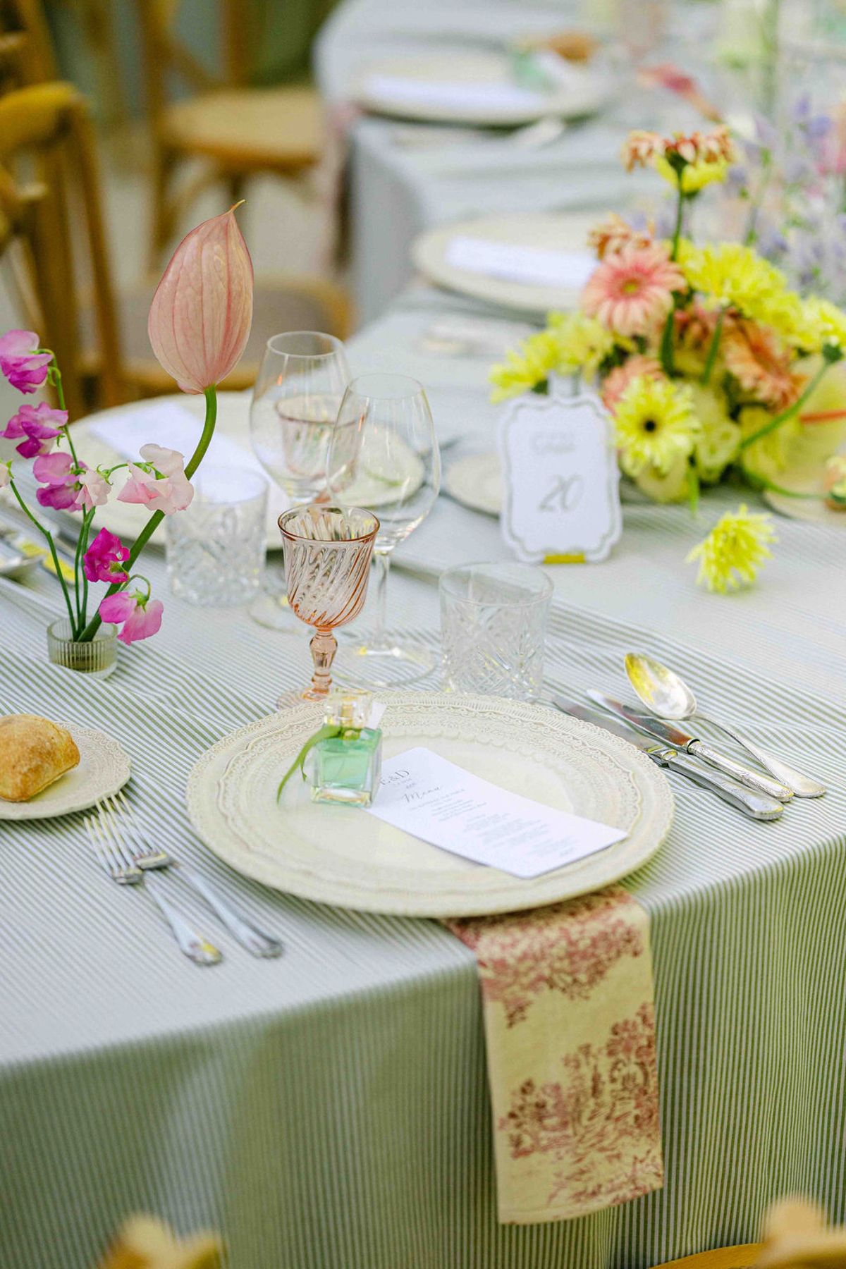 A round table laid with a striped cloth, pink glassware, a menu on a plate and a bright bouquet of yellow and pink flowers.
