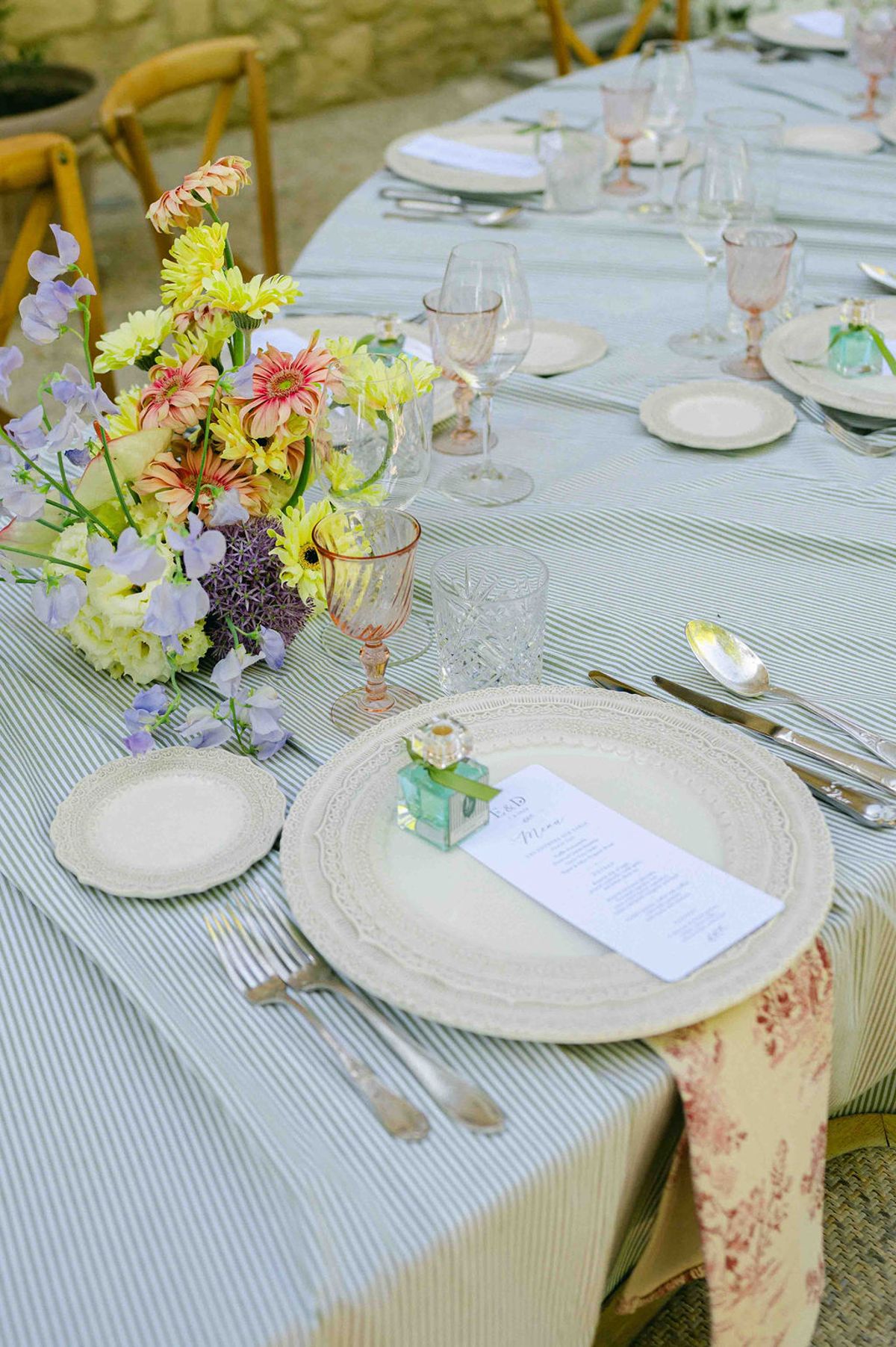 A loose centrepiece of yellow, pink and purple flowers sits beside plates, glasses and a small green bottle on a striped cloth.