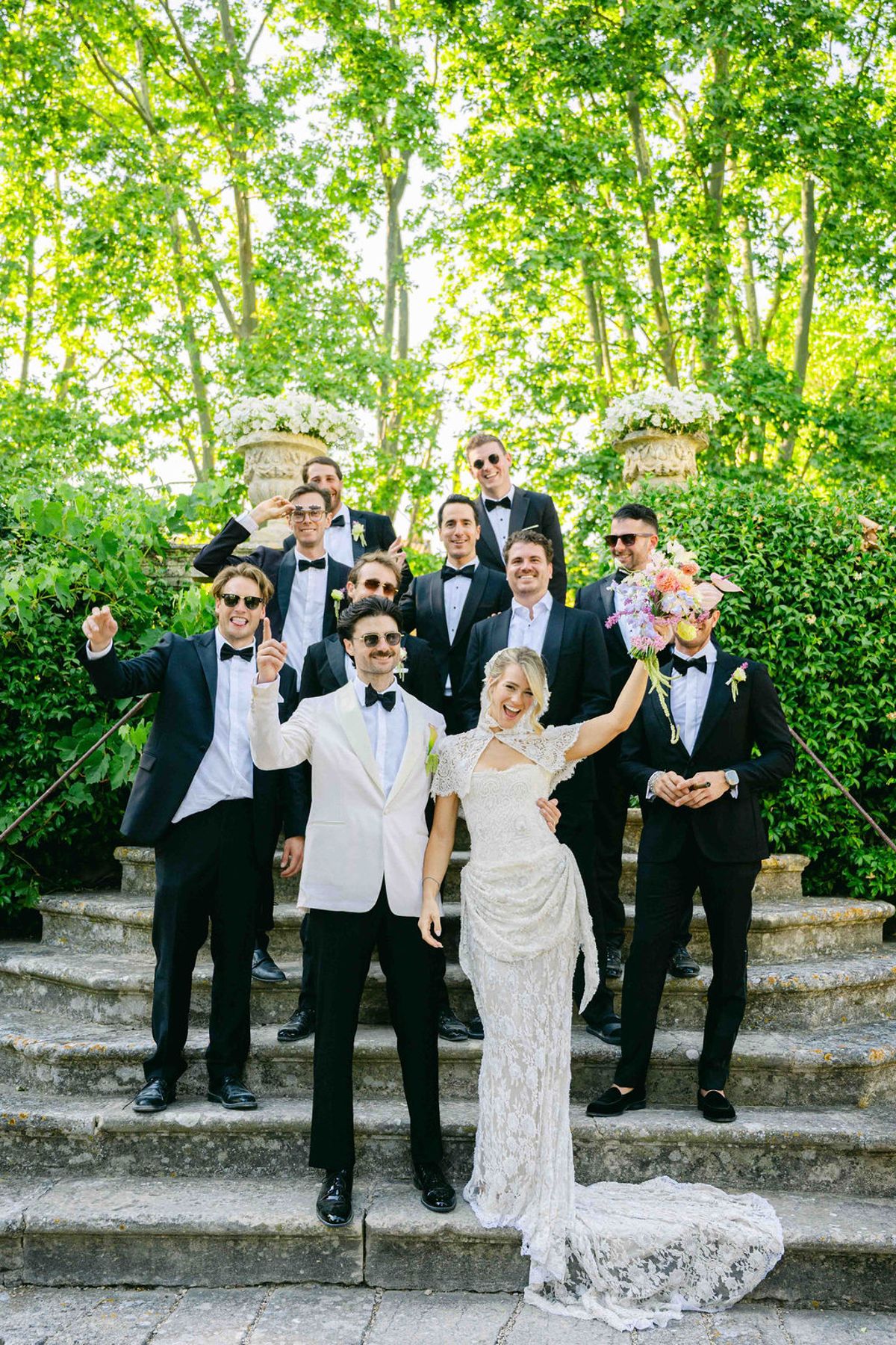 The groom in a white jacket and the bride in a lace gown pose cheerfully with a group of men in dark suits on stone steps.