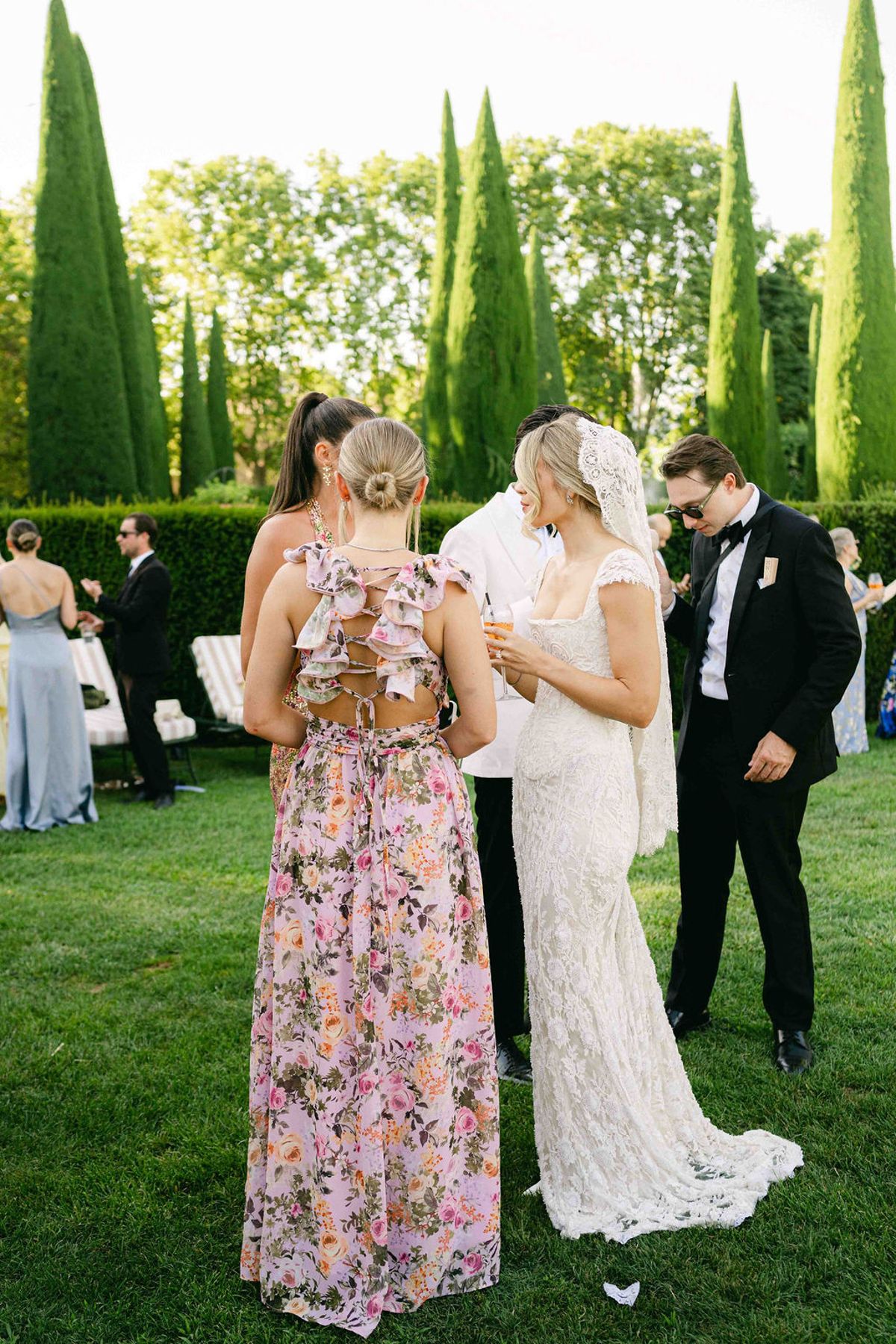 The bride and groom stand on a lawn talking with a woman in a pink floral dress and another guest.