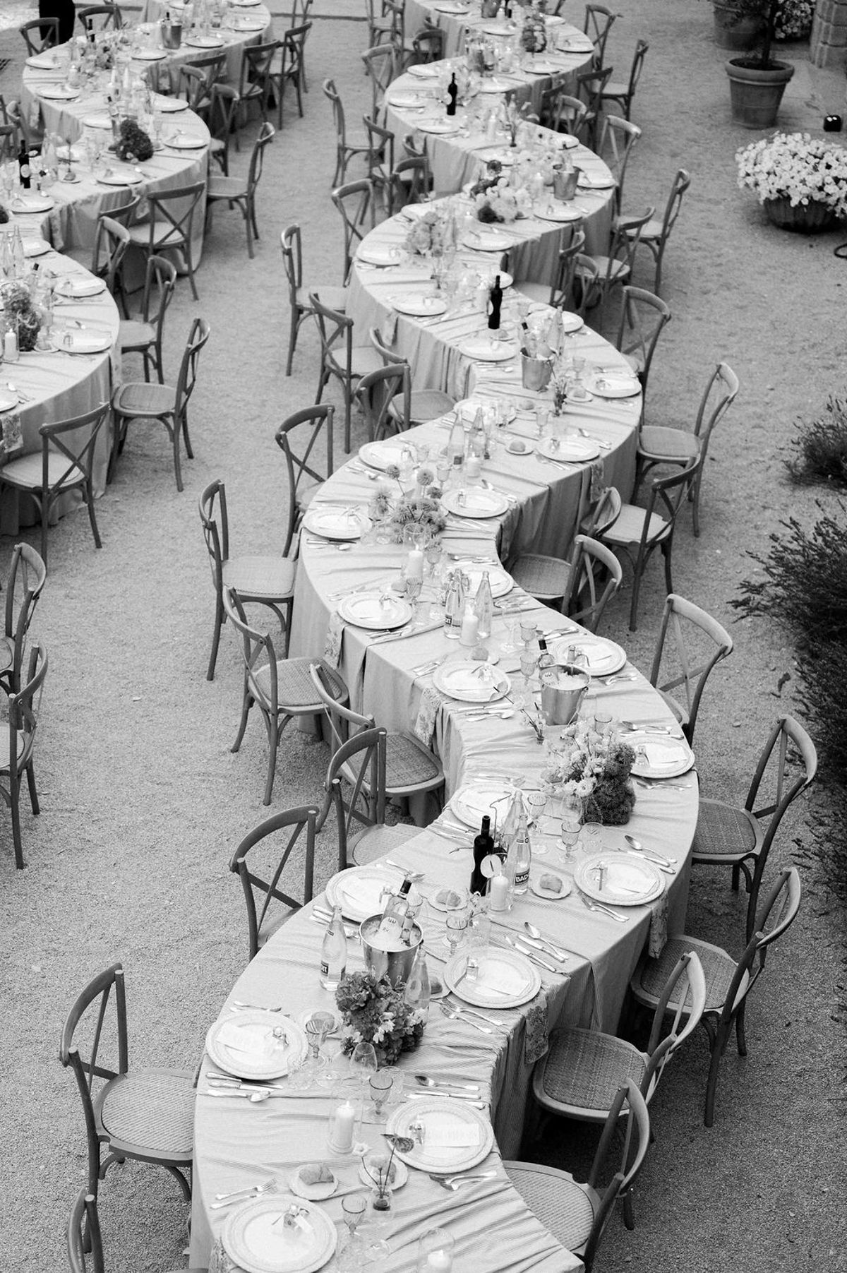 In black and white, a winding serpentine banquet table with place settings and wooden chairs is seen from above on a gravel courtyard.
