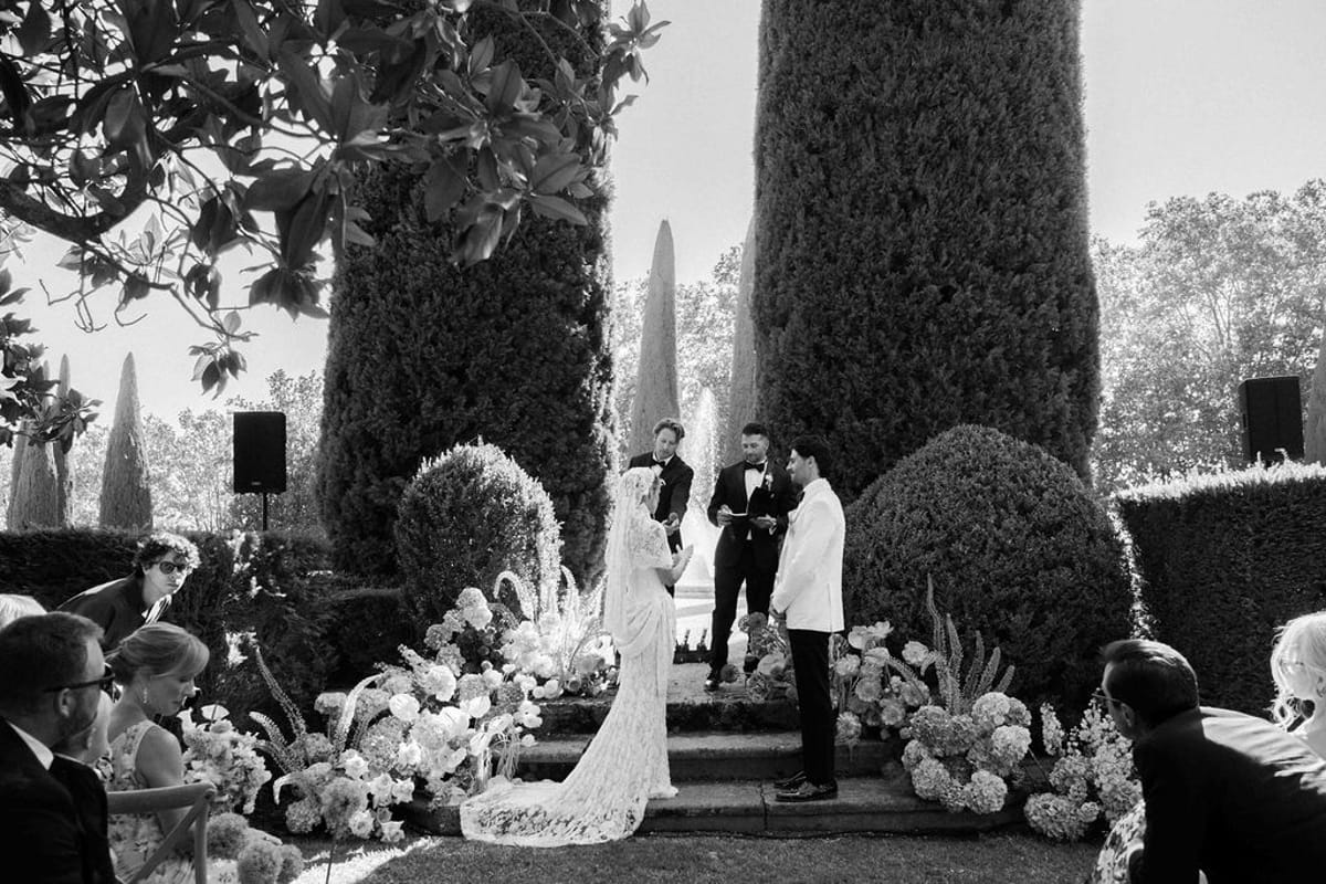In black and white, the couple and an officiant stand on flower-lined steps between cypress trees while guests watch.