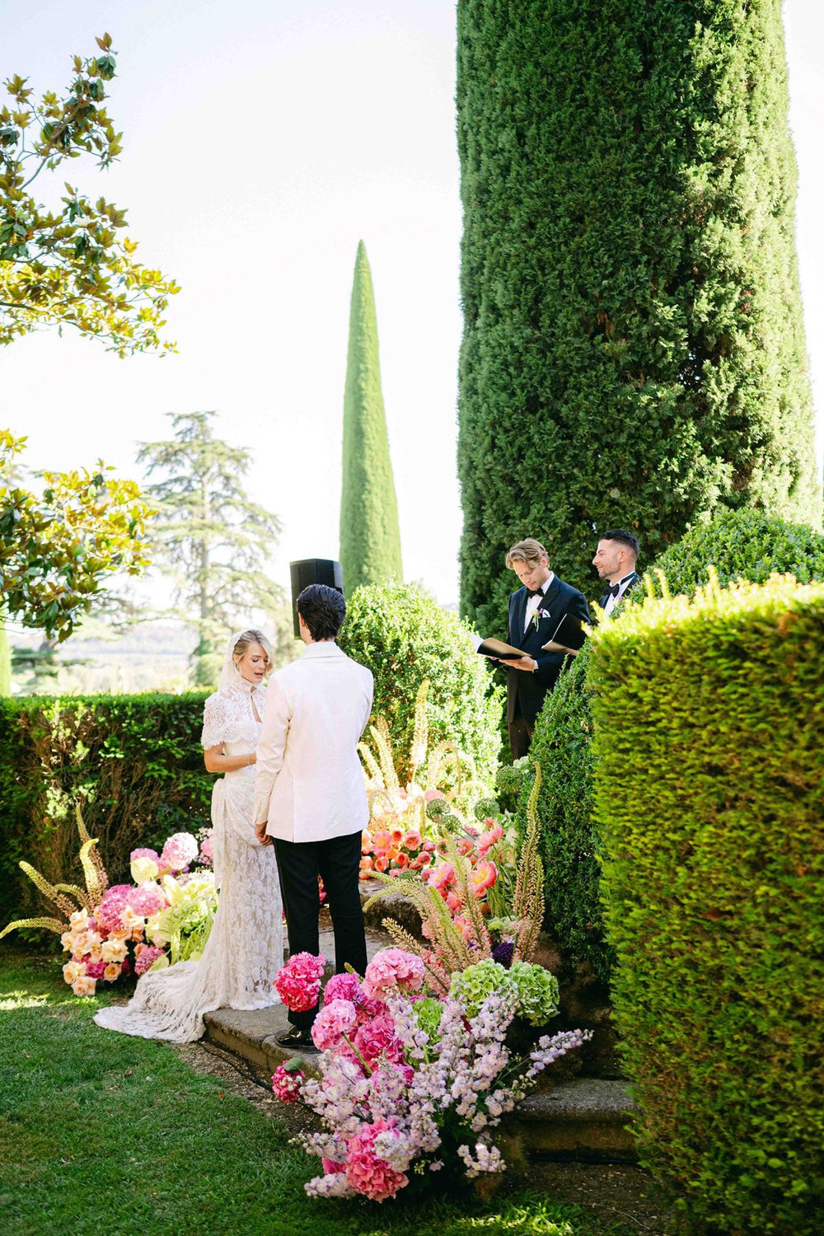 The couple stand at an altar framed by pink flowers and tall hedges, with an officiant and another person nearby.