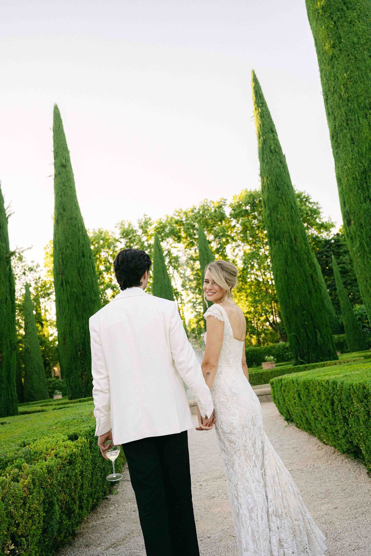 The couple walk hand in hand away from the camera along a gravel path lined with clipped hedges and tall cypress trees.