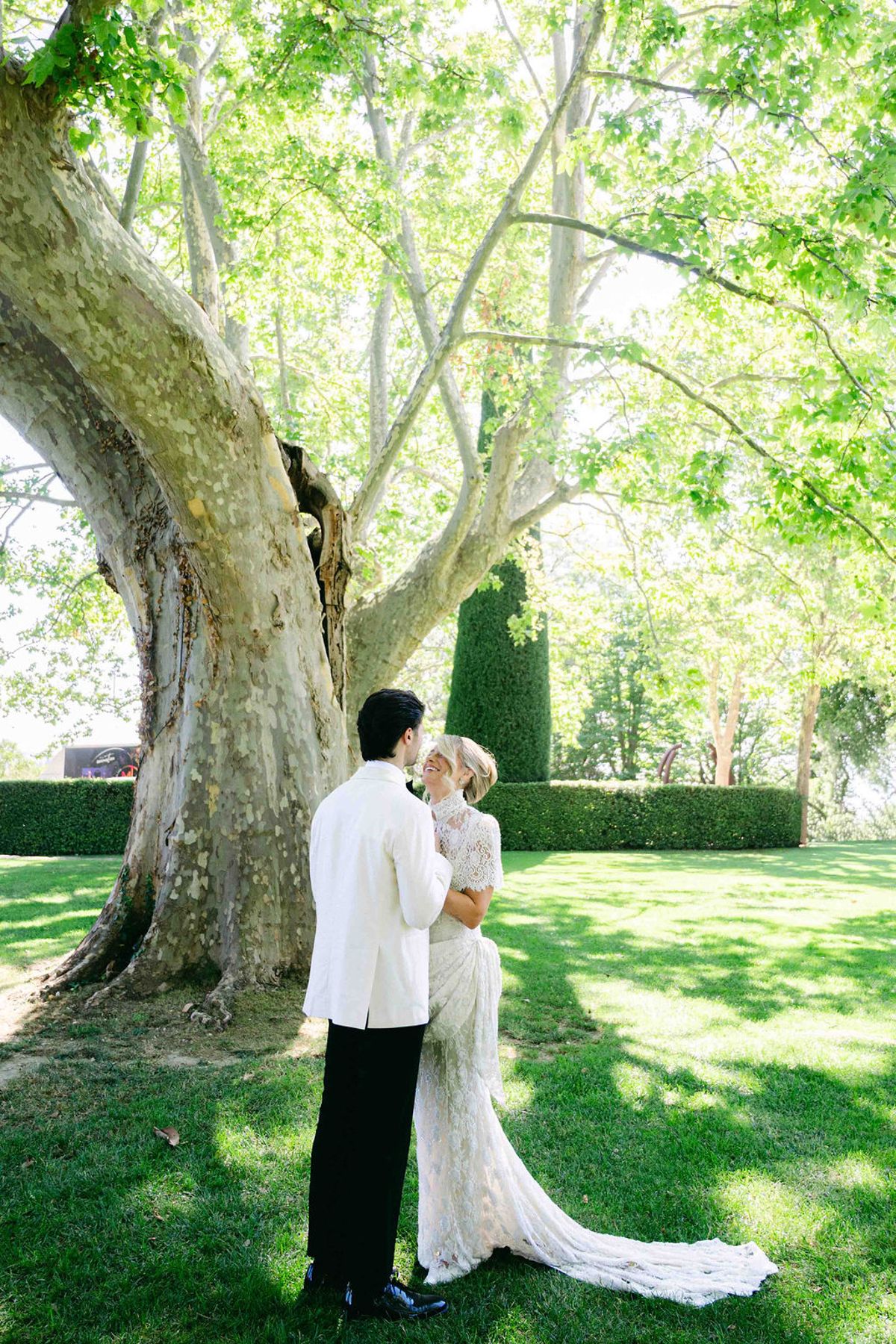 The groom in a white jacket holds the bride close on a sunlit lawn beneath a large tree with green foliage.