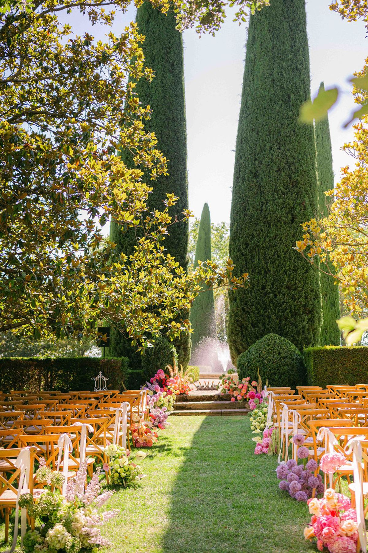 A grass aisle lined with wooden chairs and pink flowers leads towards steps and tall cypress trees in a garden.