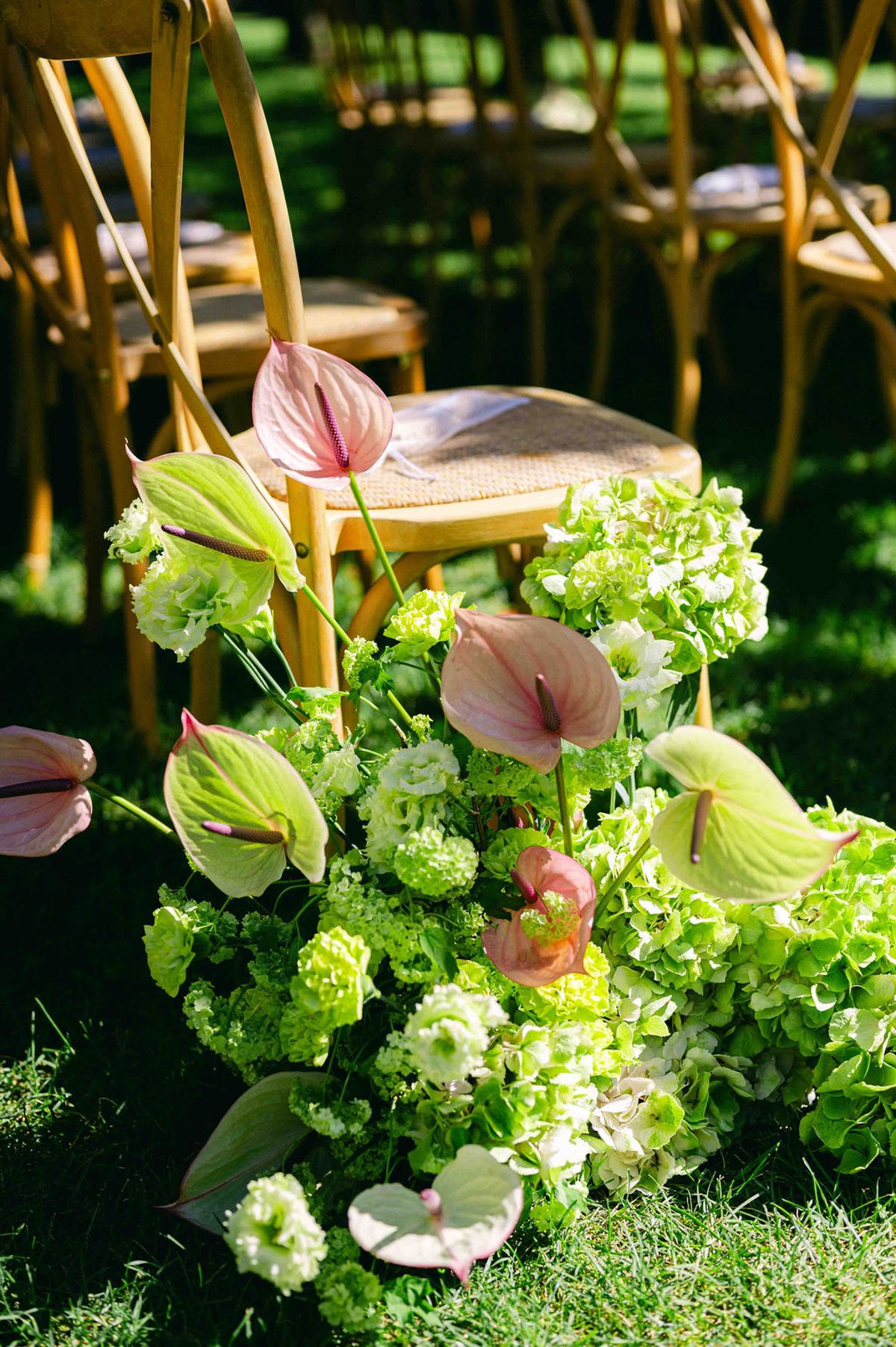 A tall arrangement of pink anthuriums and green hydrangeas stands on grass beside wooden cross-back chairs in dappled sunlight.