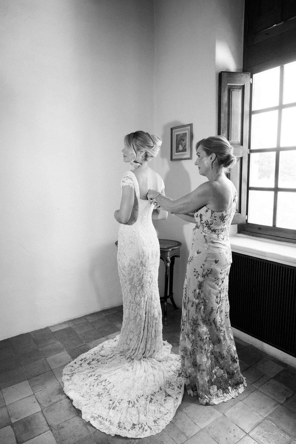In black and white, a woman fastens the back of the bride's lace gown beside a bright window.