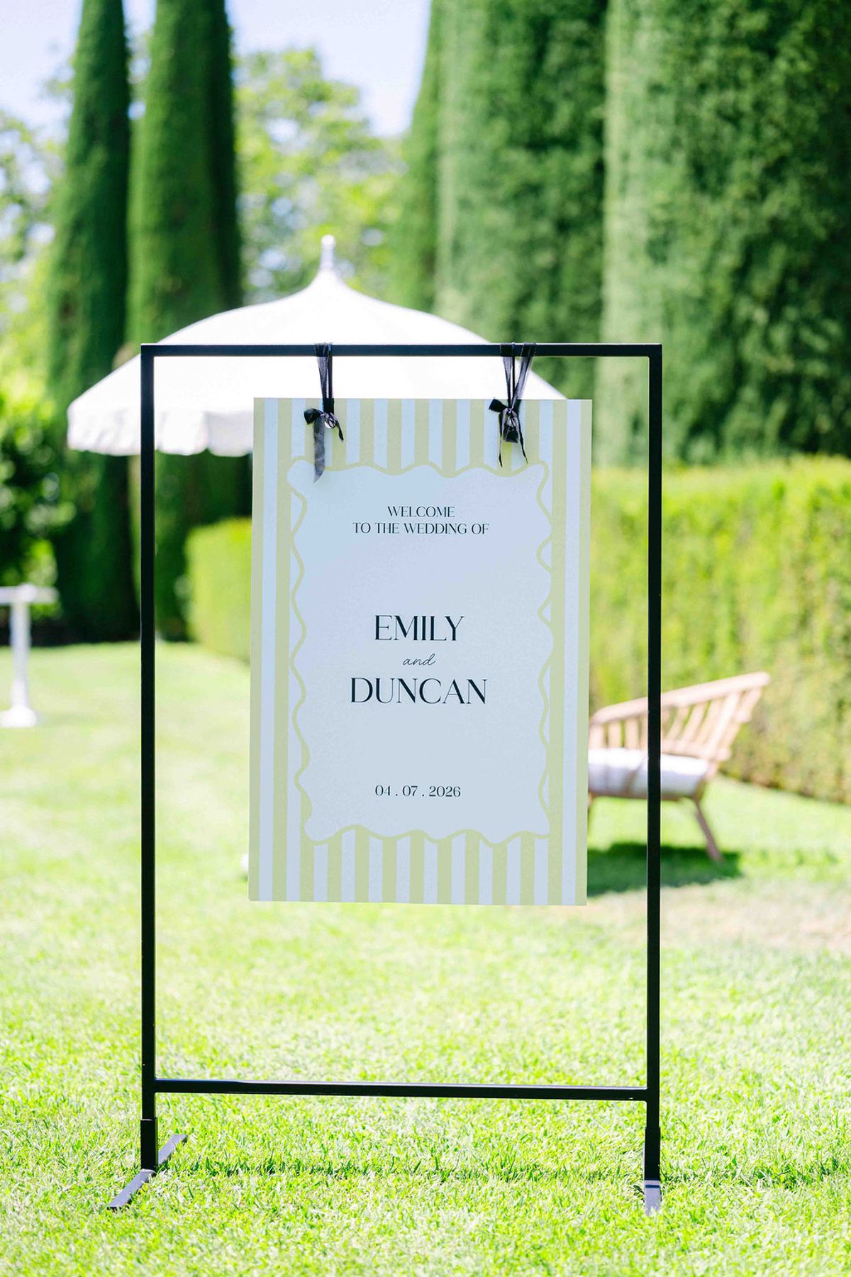 A striped welcome sign hangs from a black frame on a lawn beside a white parasol, with tall cypress hedges behind.
