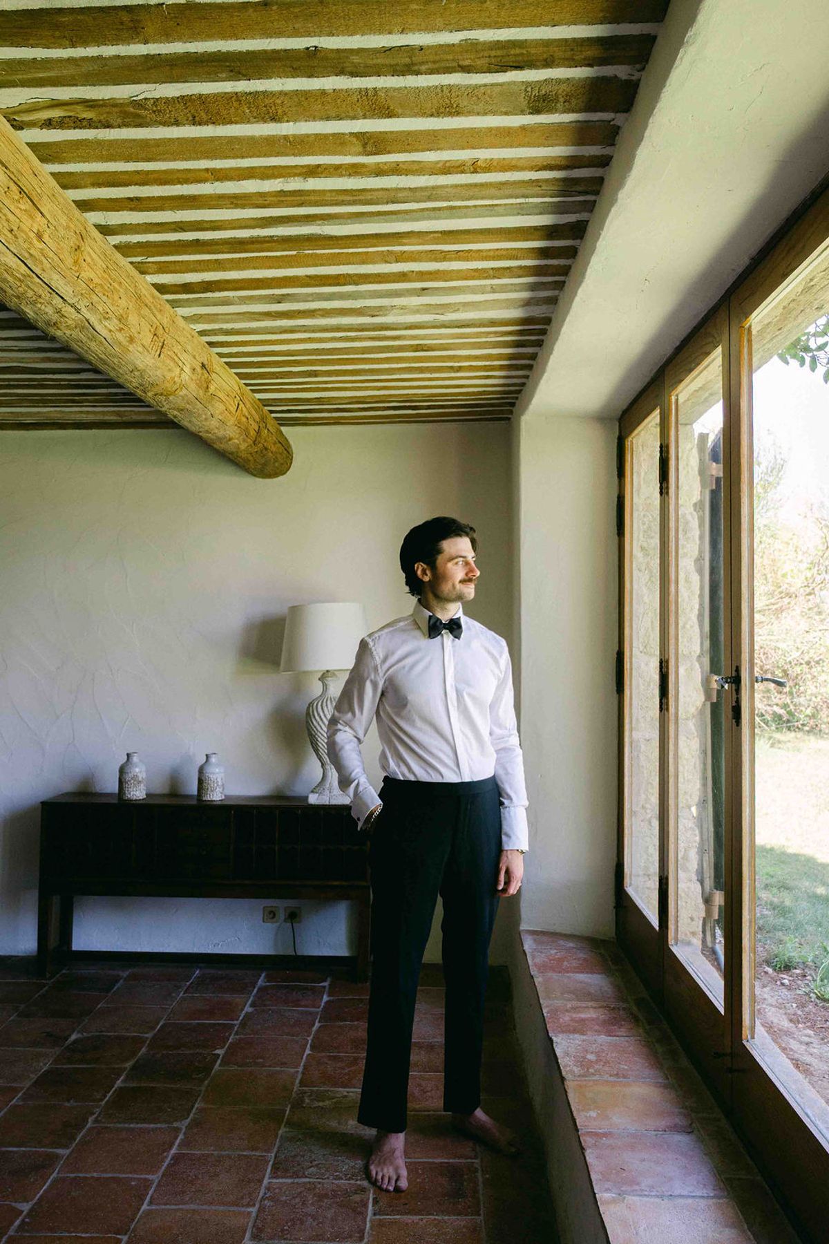 The groom in a white shirt, black trousers and bow tie stands barefoot beside a large window in a room with a beamed ceiling.