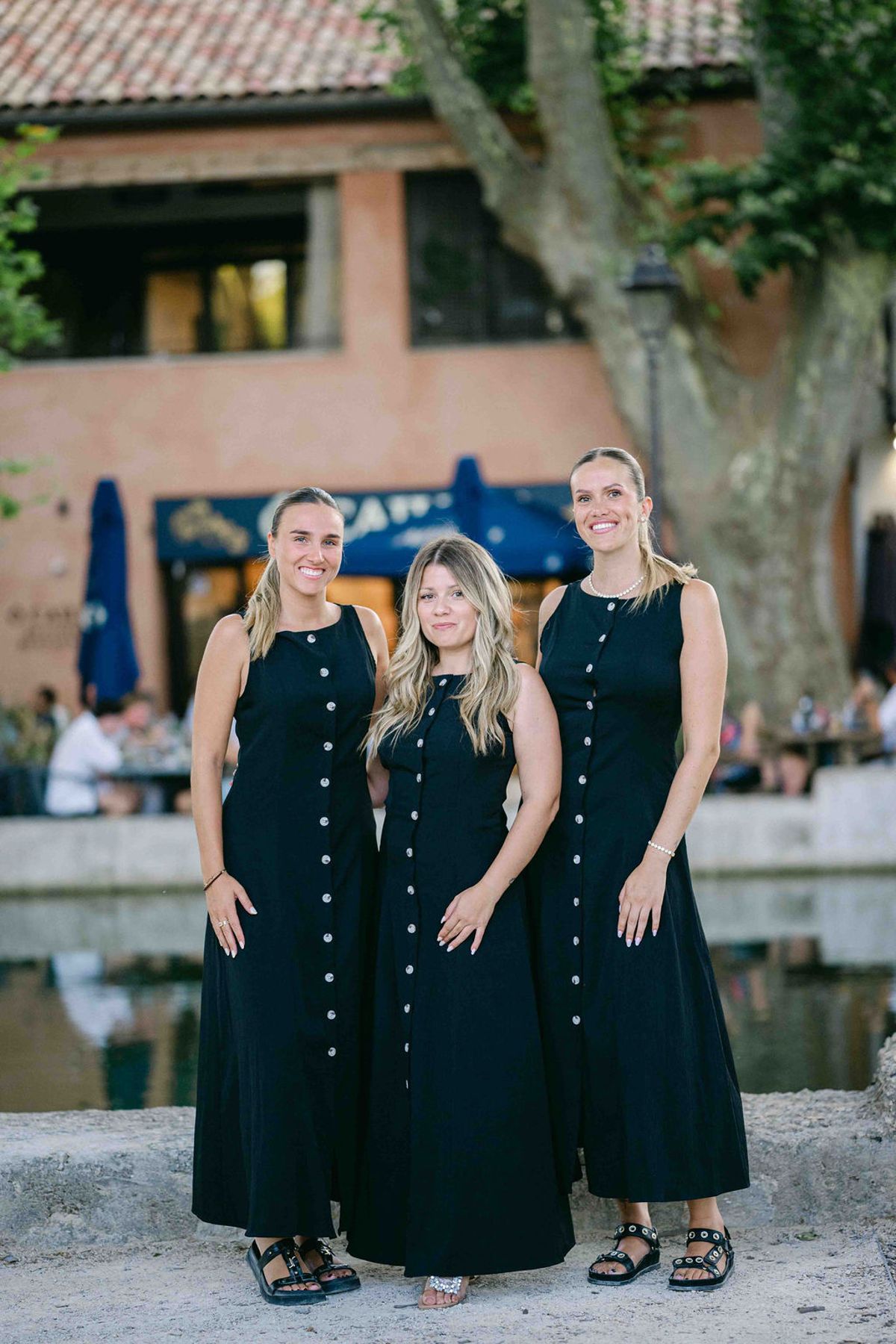 Three smiling women in long navy buttoned dresses stand together beside a pool of water, with a building and tree behind.