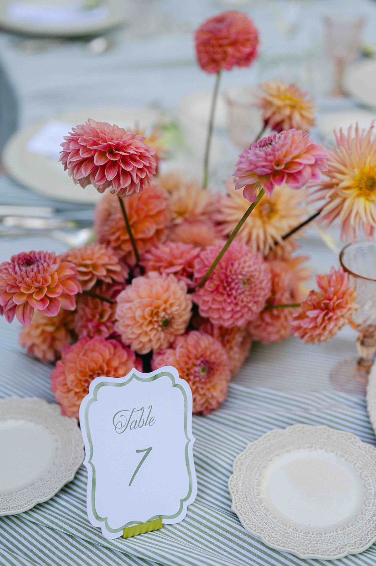 A centrepiece of orange and coral dahlias sits beside a numbered table card on a striped tablecloth with scalloped plates.
