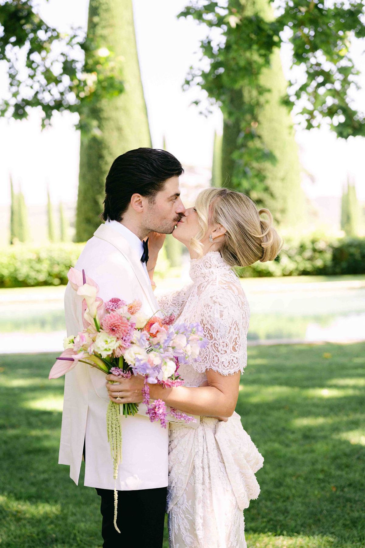 The groom in a white jacket kisses the bride in lace while holding a pastel bouquet, on a lawn with tall trees.