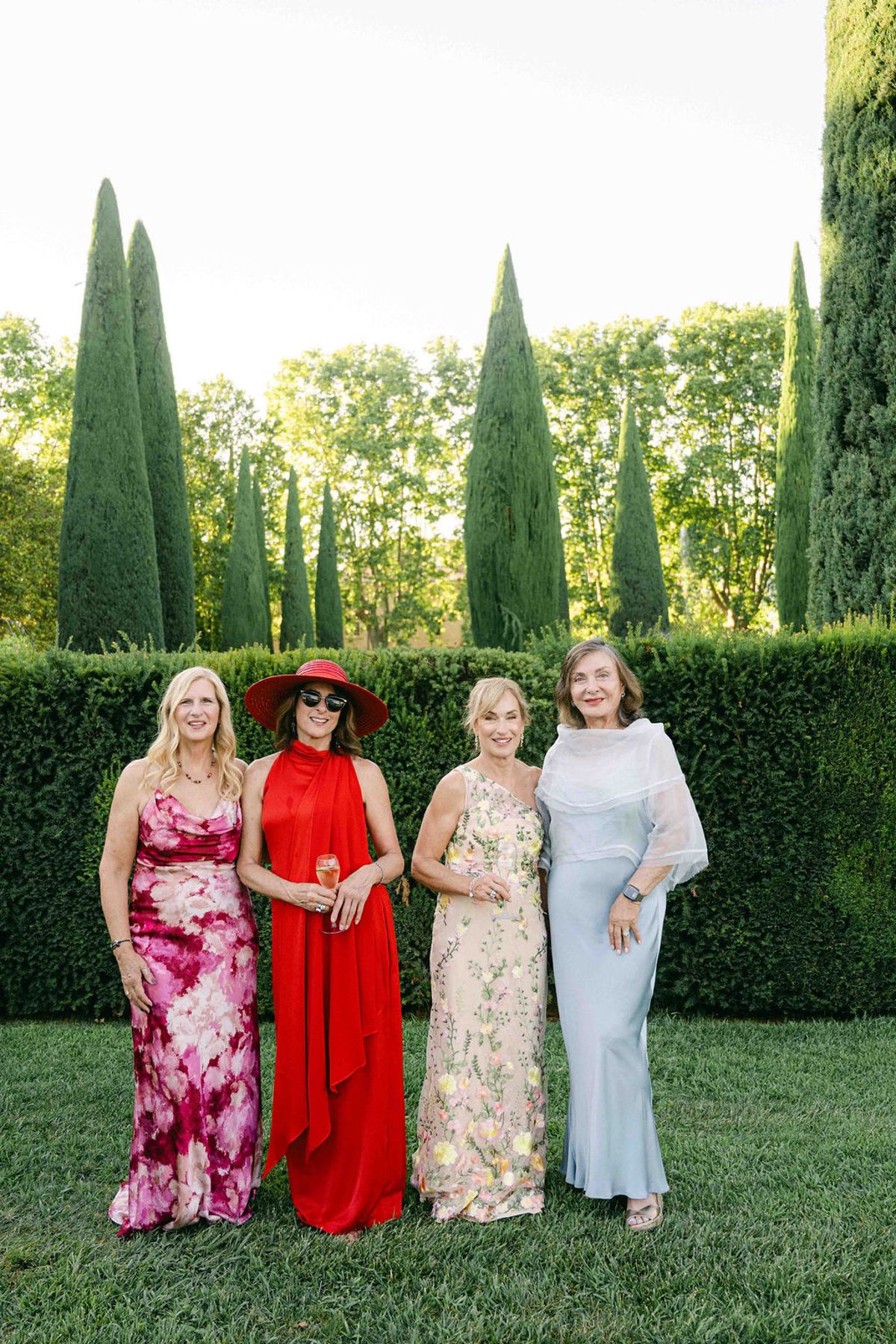 Four women in pink floral, red, yellow floral and pale blue dresses stand together on a lawn in front of a tall clipped hedge.