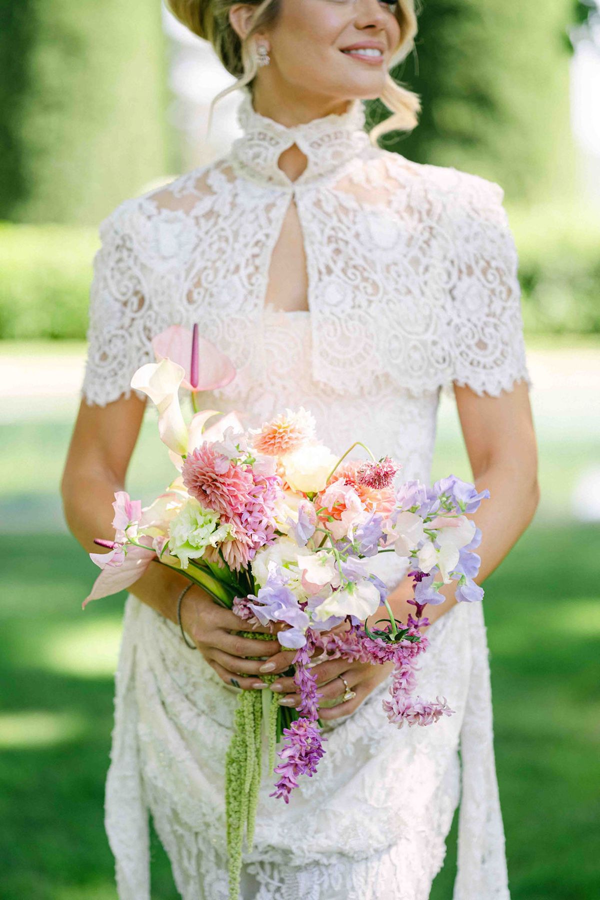 The bride in a high-collared lace dress smiles while holding a bouquet of pink and lilac flowers on a green lawn.