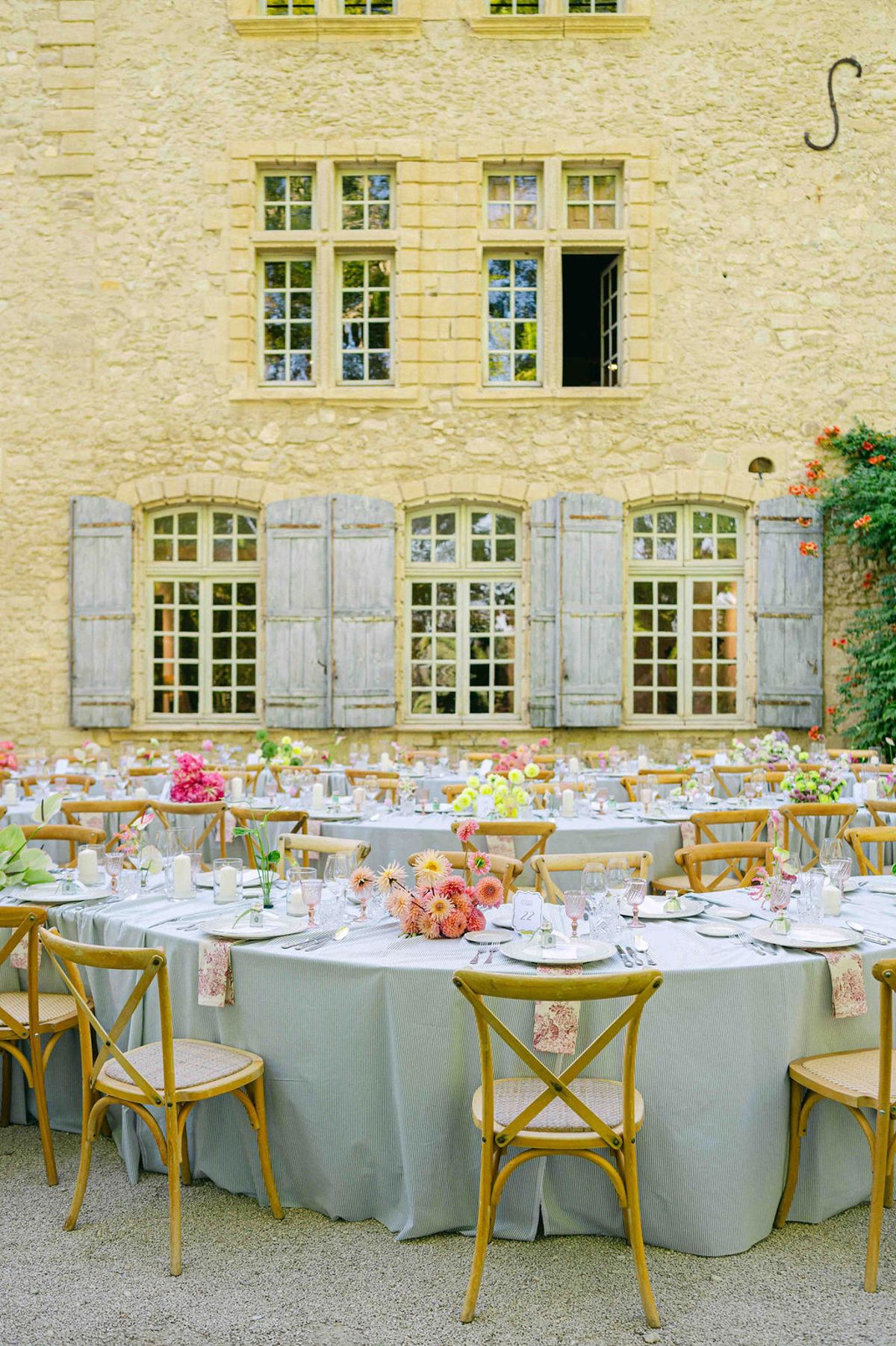Round tables with grey linen, pastel flower centrepieces and wooden cross-back chairs stand on gravel before a pale stone chateau with grey shutters.