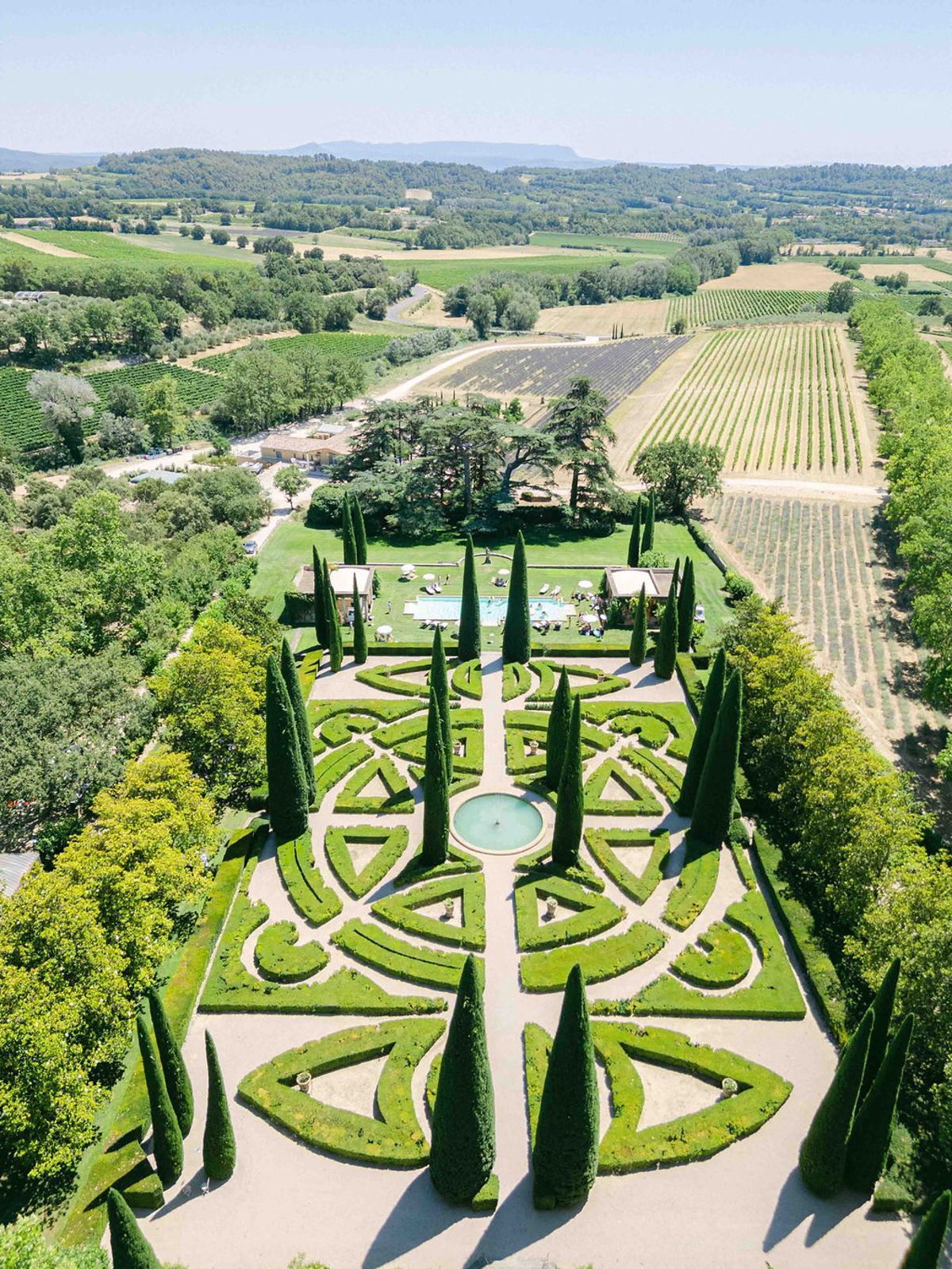 An aerial view of a formal knot garden with clipped hedges and cypress trees, with vineyards and hills beyond.