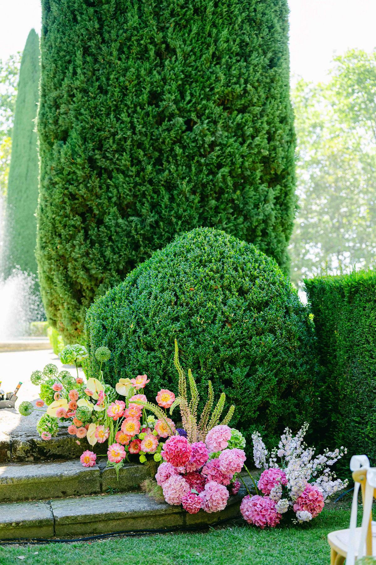 Pink and orange hydrangeas and other flowers sit beside stone steps, below a clipped round topiary and a tall hedge.