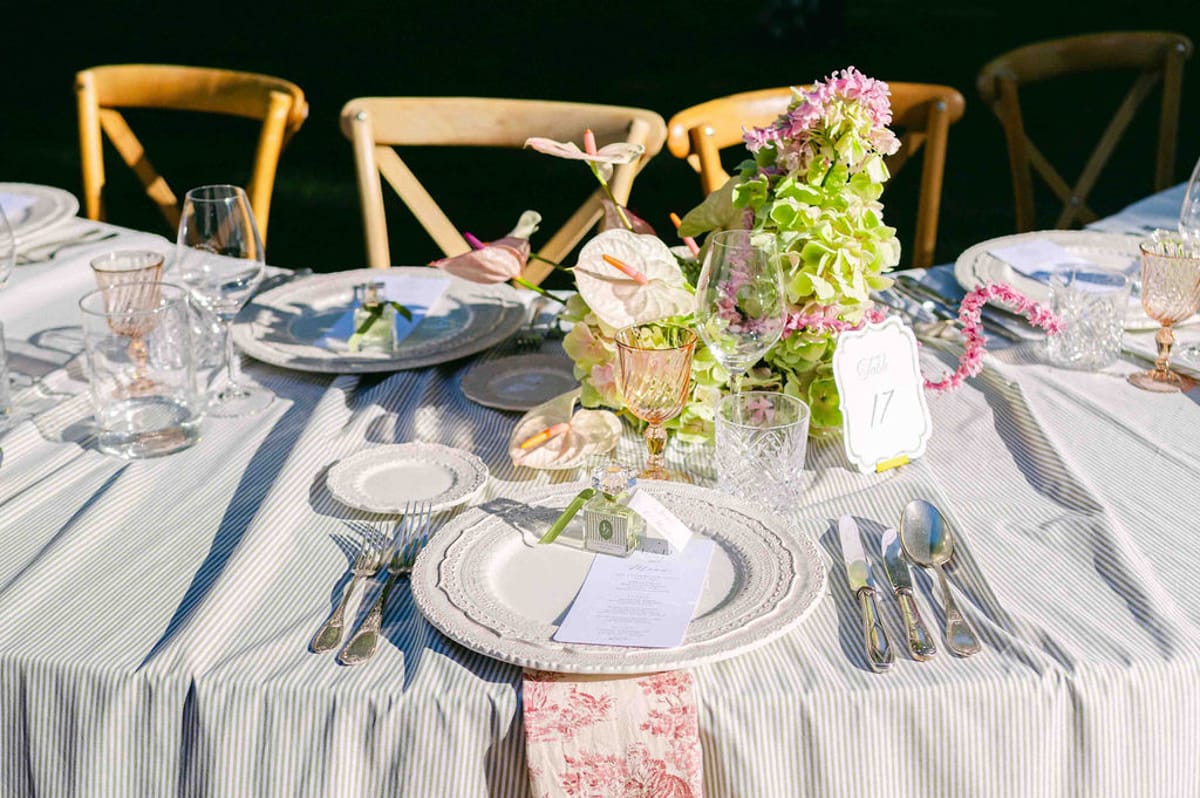 A table laid with a striped blue and white cloth, a plate with a menu card, glassware and a colourful floral centrepiece.