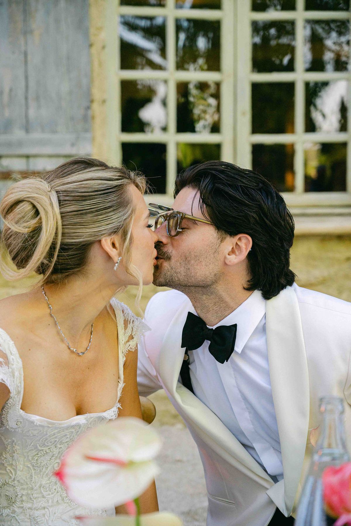 The groom in a white dinner jacket and sunglasses kisses the bride beside a tall stone window with shutters.