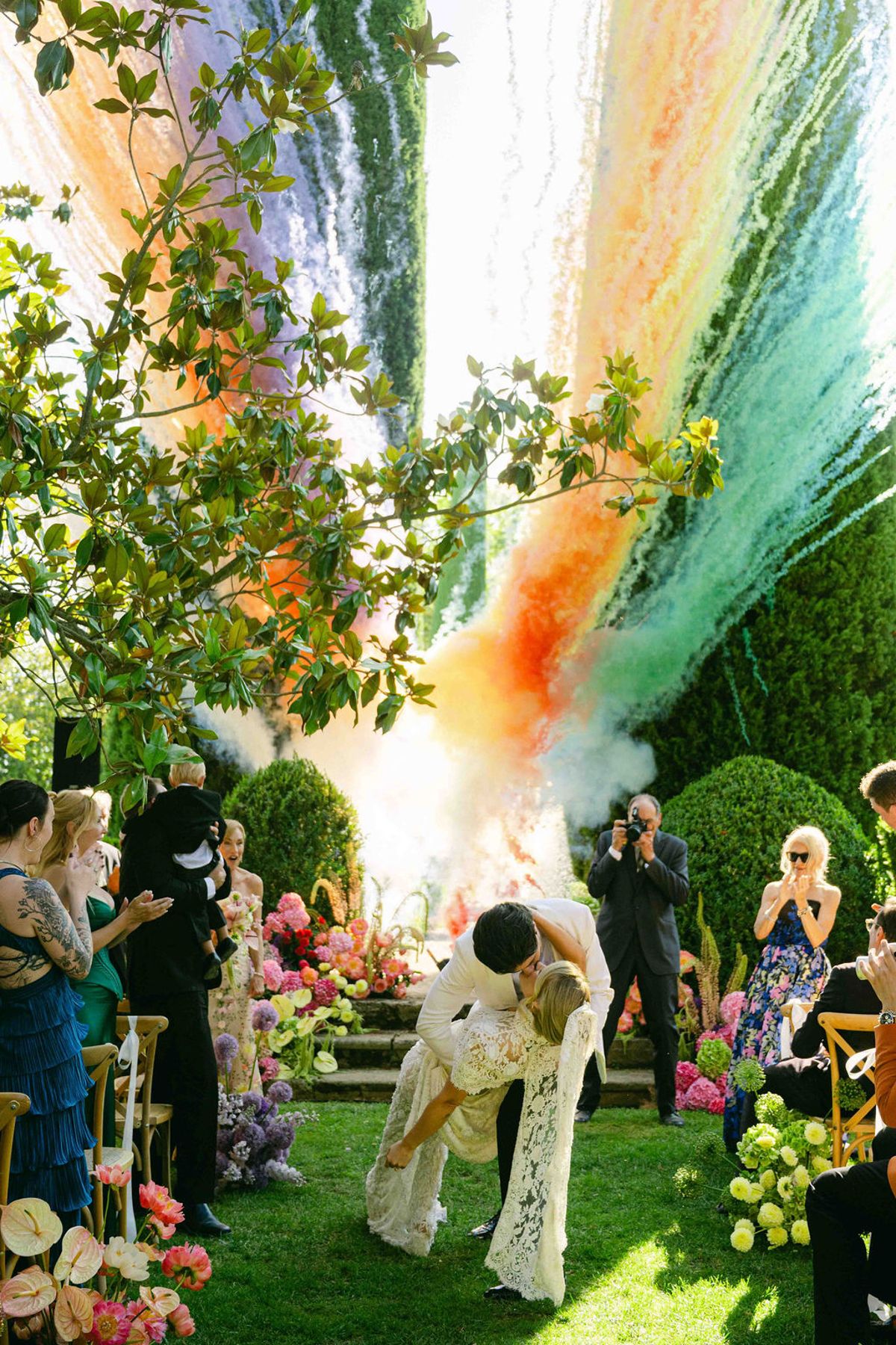 The couple kiss in an aisle lined with flowers as orange, green and blue smoke drifts overhead and guests watch and photograph.