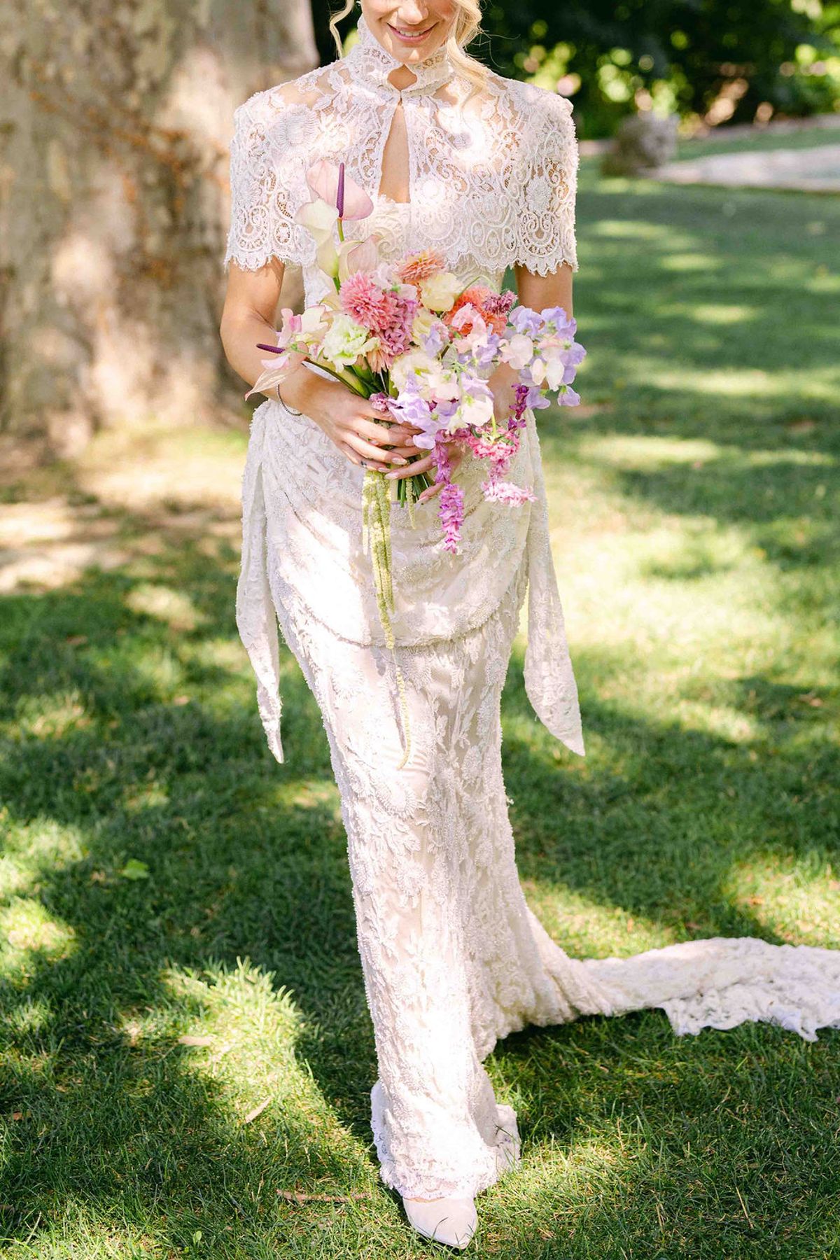 A bride in a lace jacket and long gown stands on dappled grass holding a pastel bouquet, her face out of frame.
