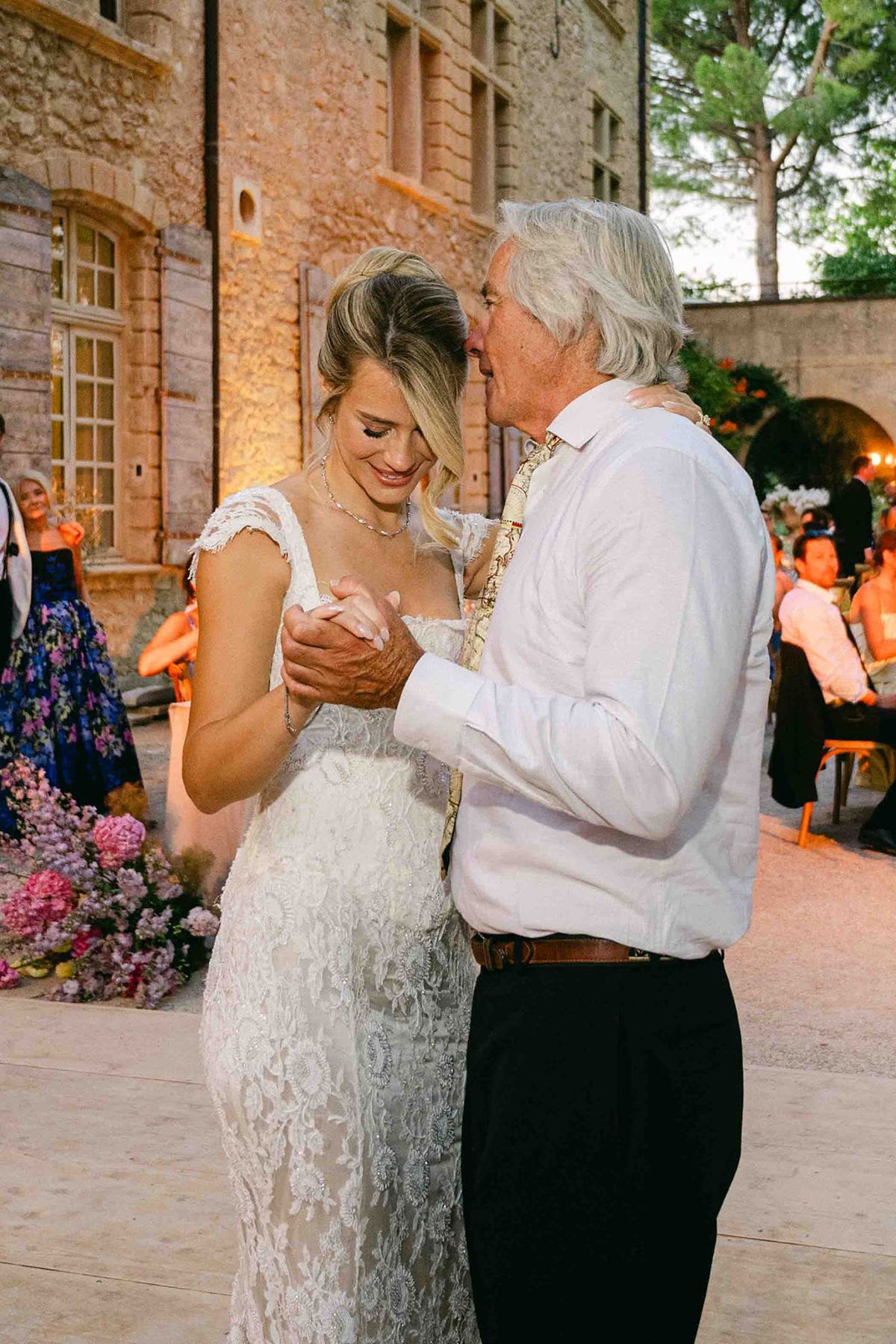 The bride in a lace dress dances cheek to cheek with an older man in a white shirt, with guests behind.