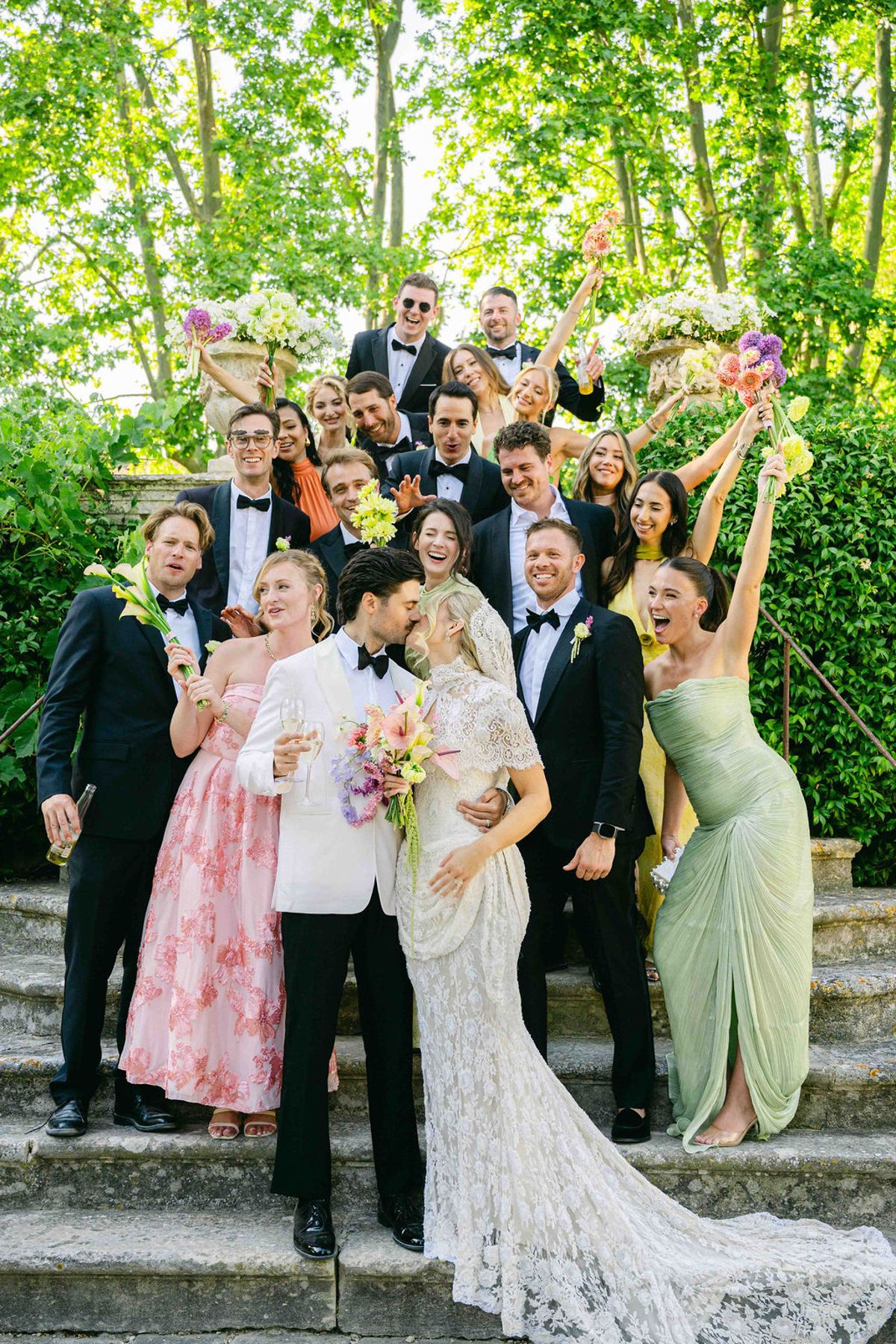 The couple and their wedding party pose cheerfully together on stone steps, some raising their arms, among green hedges.