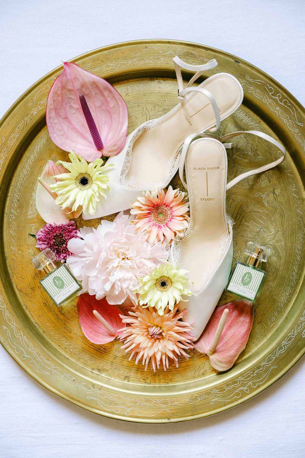 A round brass tray holds white heeled shoes, pink and orange flowers and two small perfume bottles, photographed from directly above.