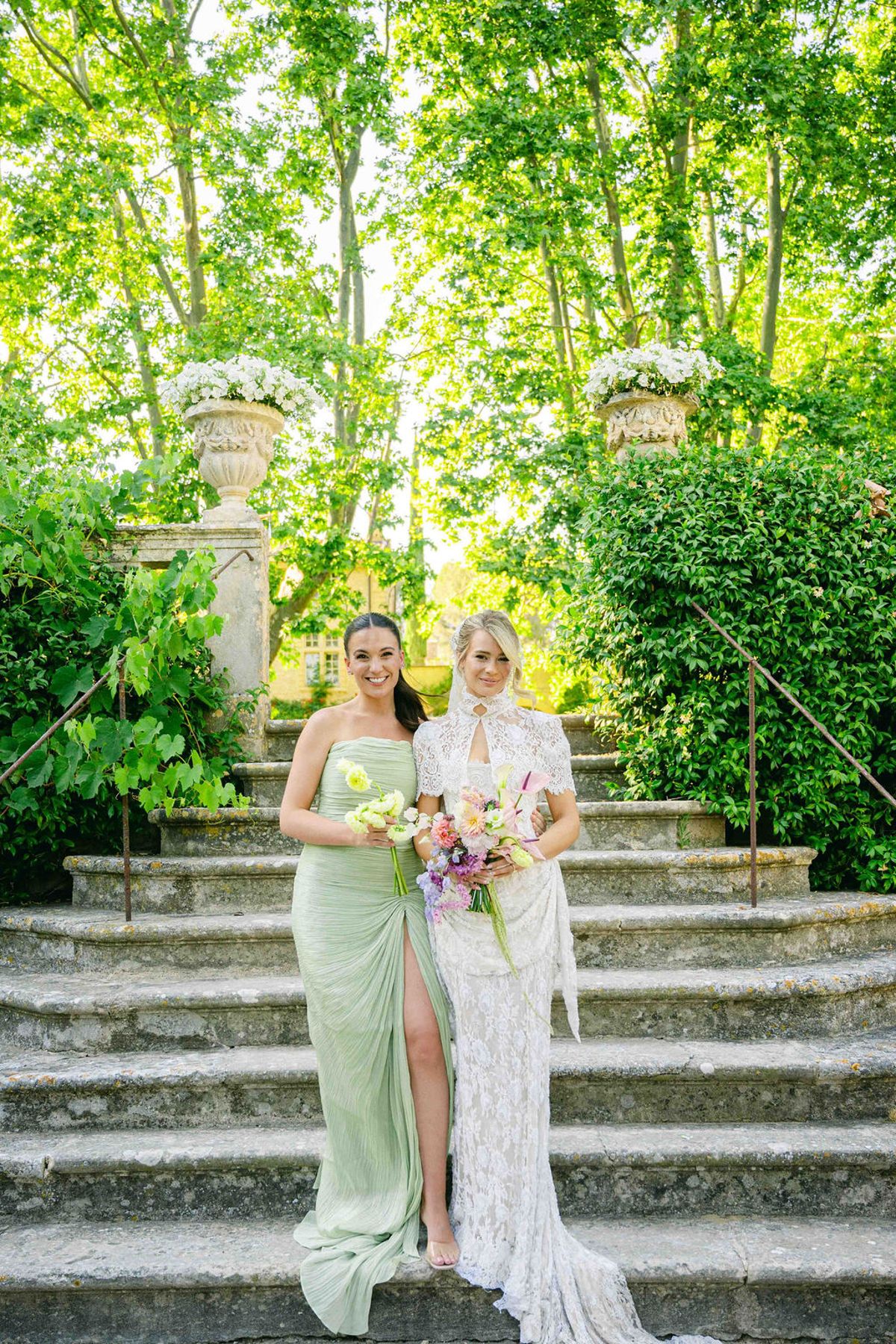 The bride in a white lace dress with a bouquet stands beside a woman in a pale green dress on stone steps.