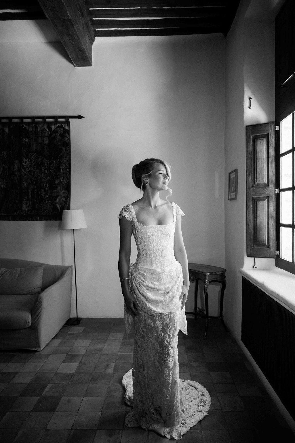 In black and white, the bride stands in her lace dress beside a window in a room with a tiled floor and beamed ceiling.