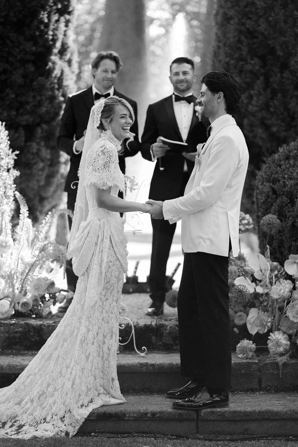 In black and white, the couple hold hands facing each other while an officiant and another man stand behind them.