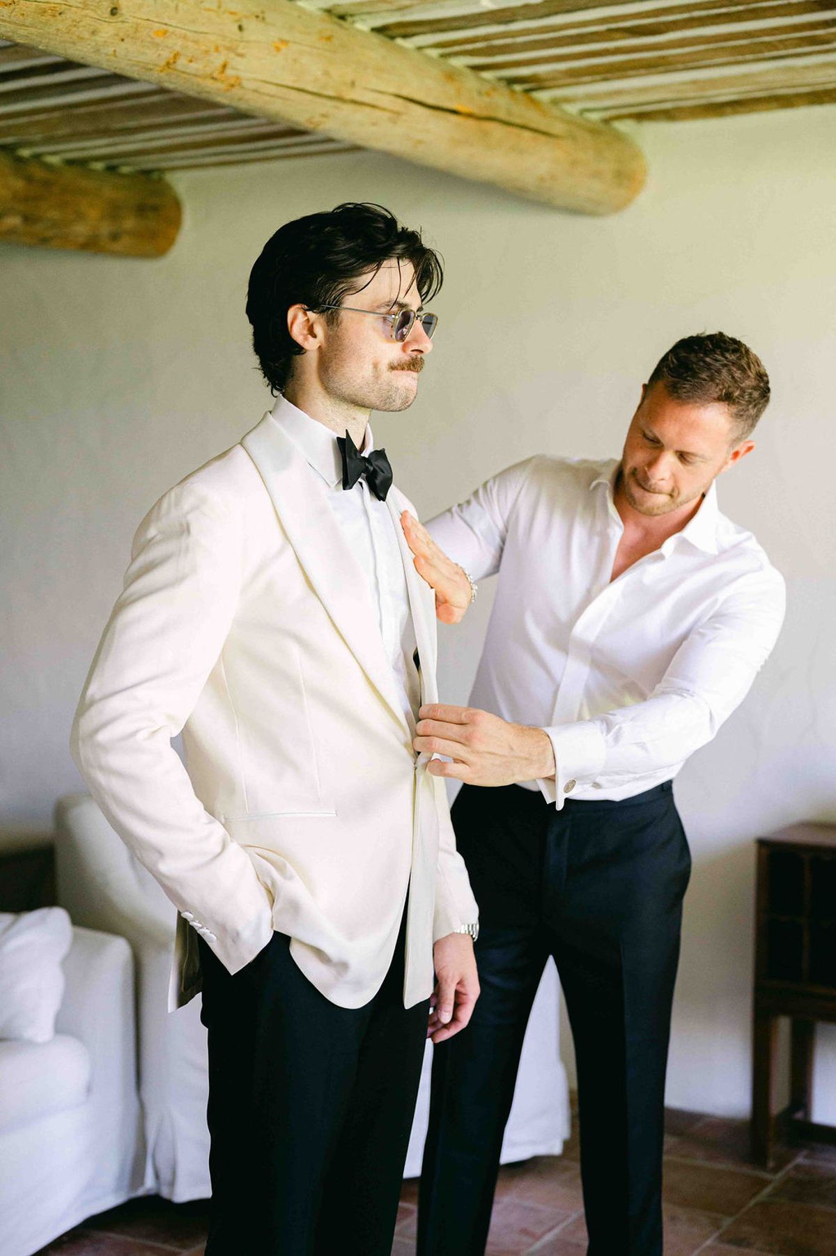 The groom in a white dinner jacket and bow tie has his lapel adjusted by a man in a white shirt.