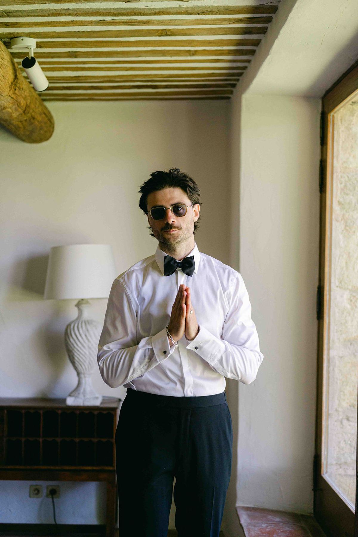 The groom in sunglasses, a white shirt and black bow tie stands indoors with his palms pressed together.