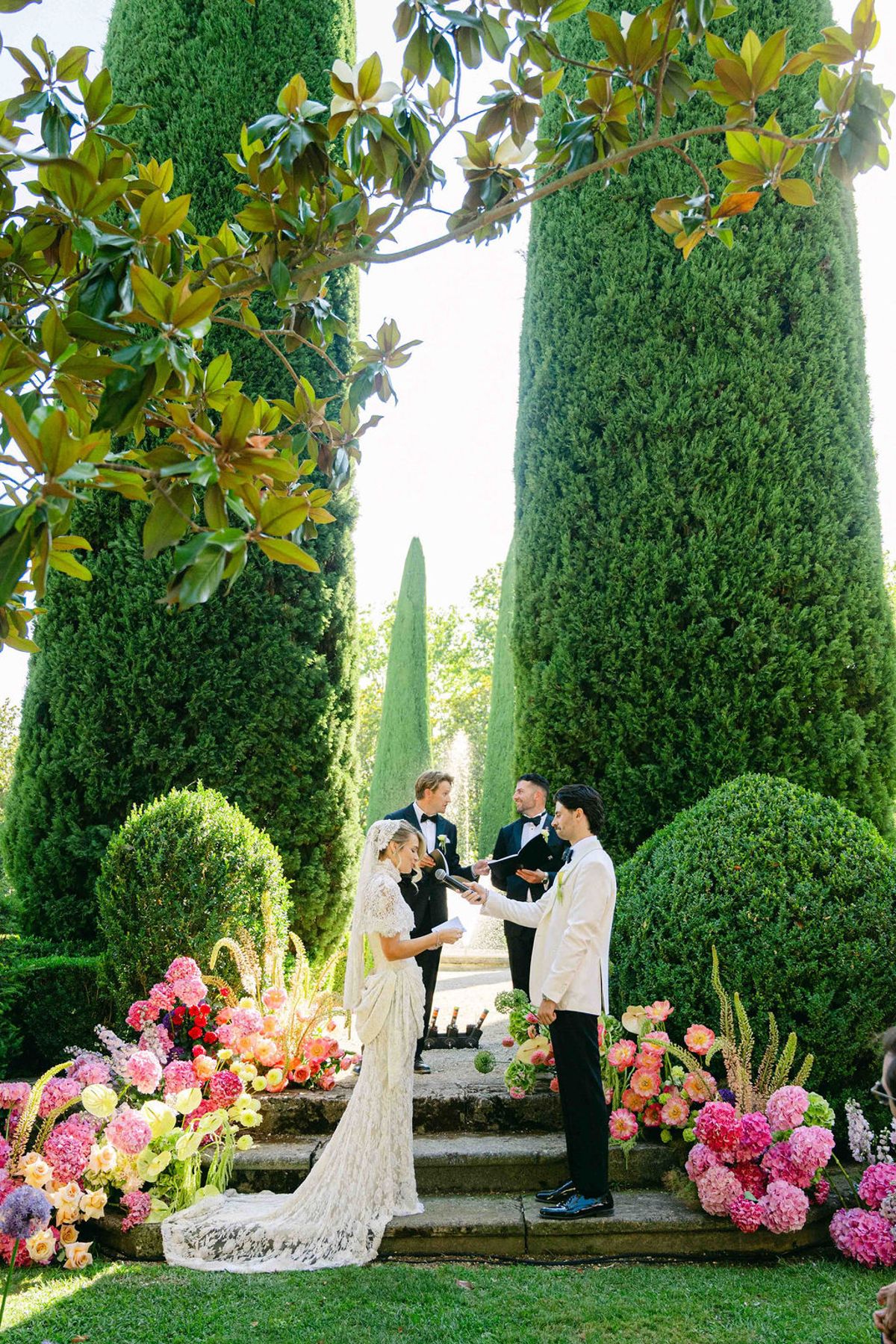The couple stand on stone steps with an officiant, surrounded by pink flowers and tall clipped cypress trees in a garden.