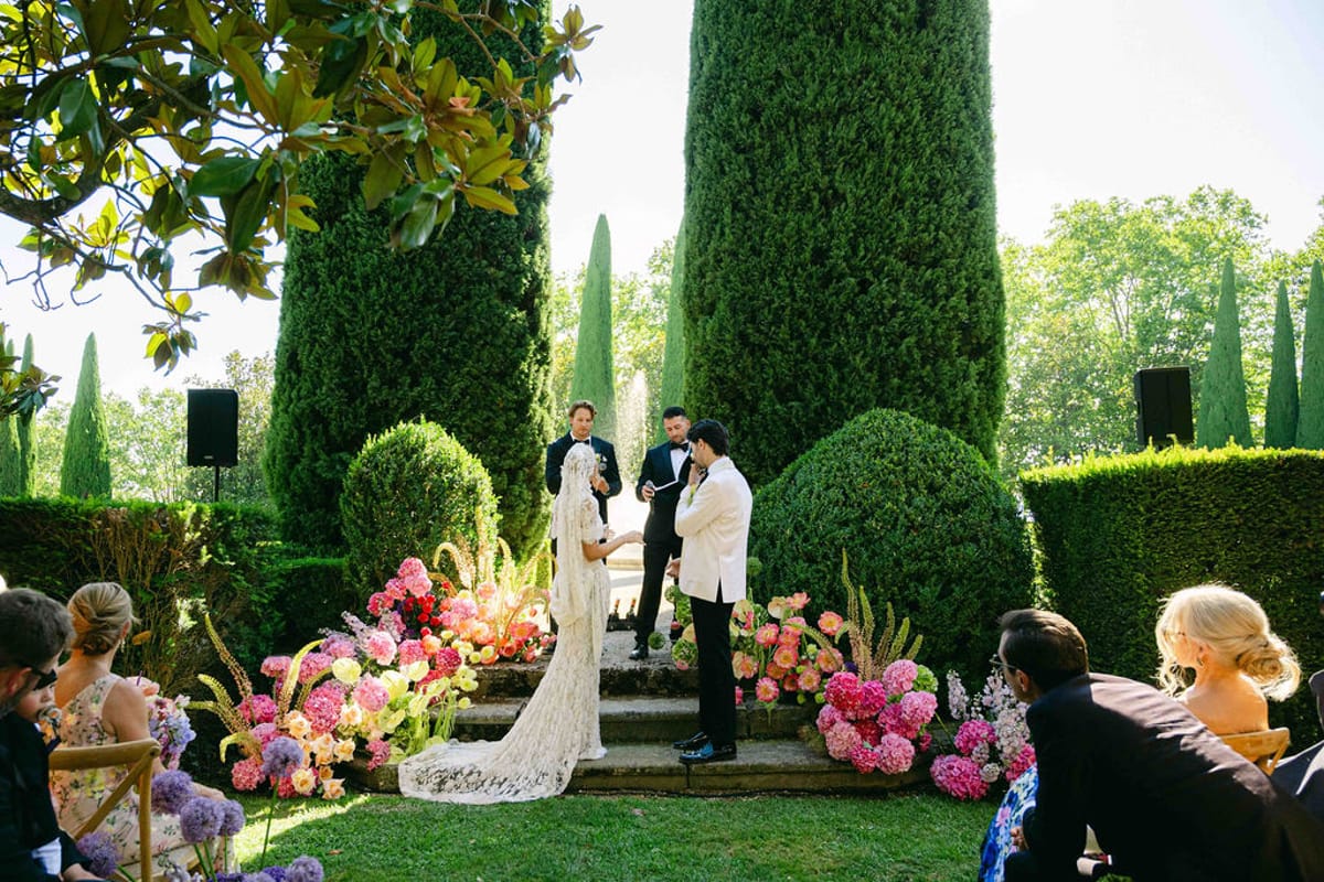 The couple face each other before an officiant on steps decorated with pink flowers, with guests seated nearby.