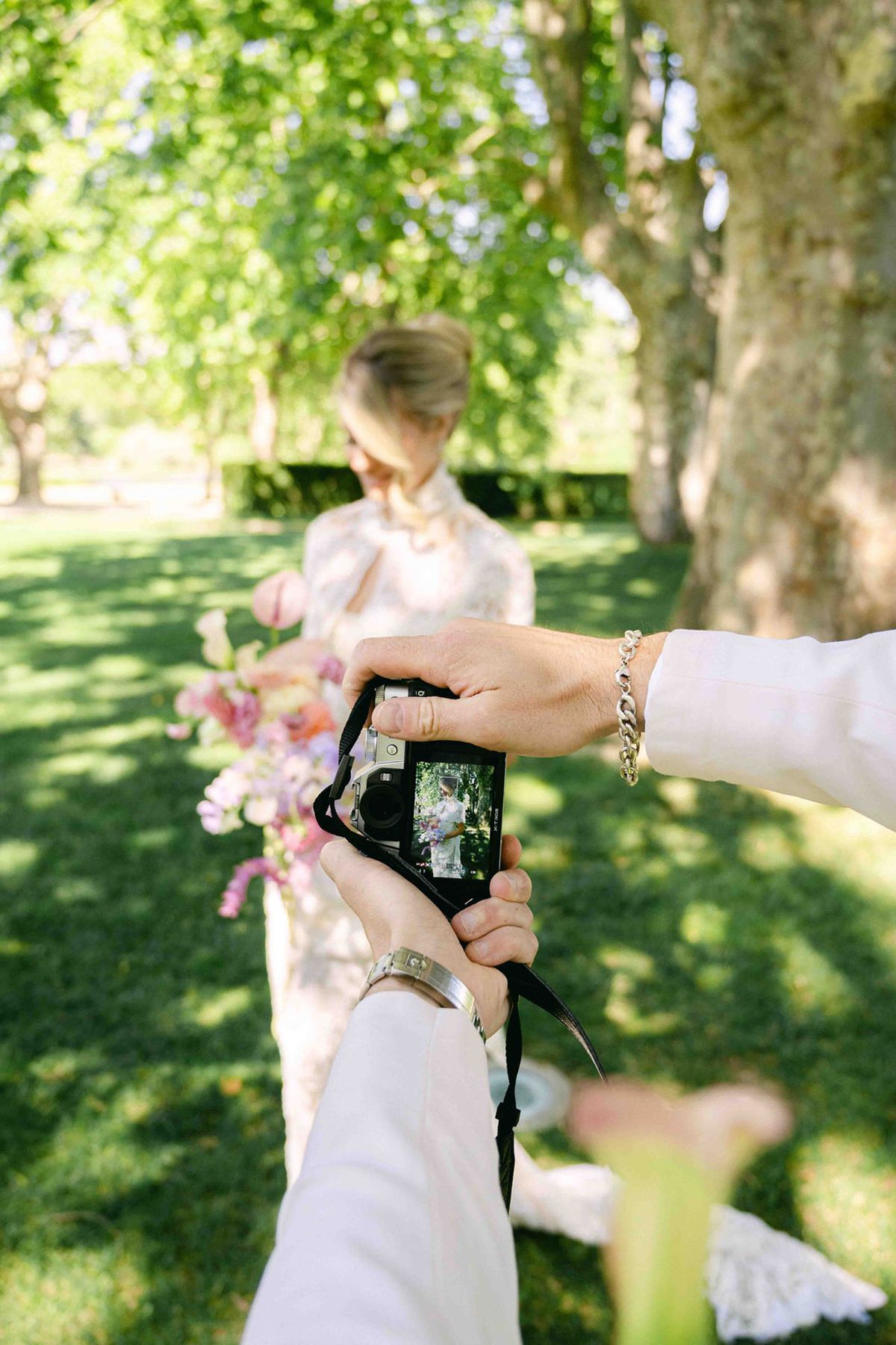 Two hands hold a camera in the foreground while the bride, holding a pastel bouquet, stands on a lawn beneath trees.