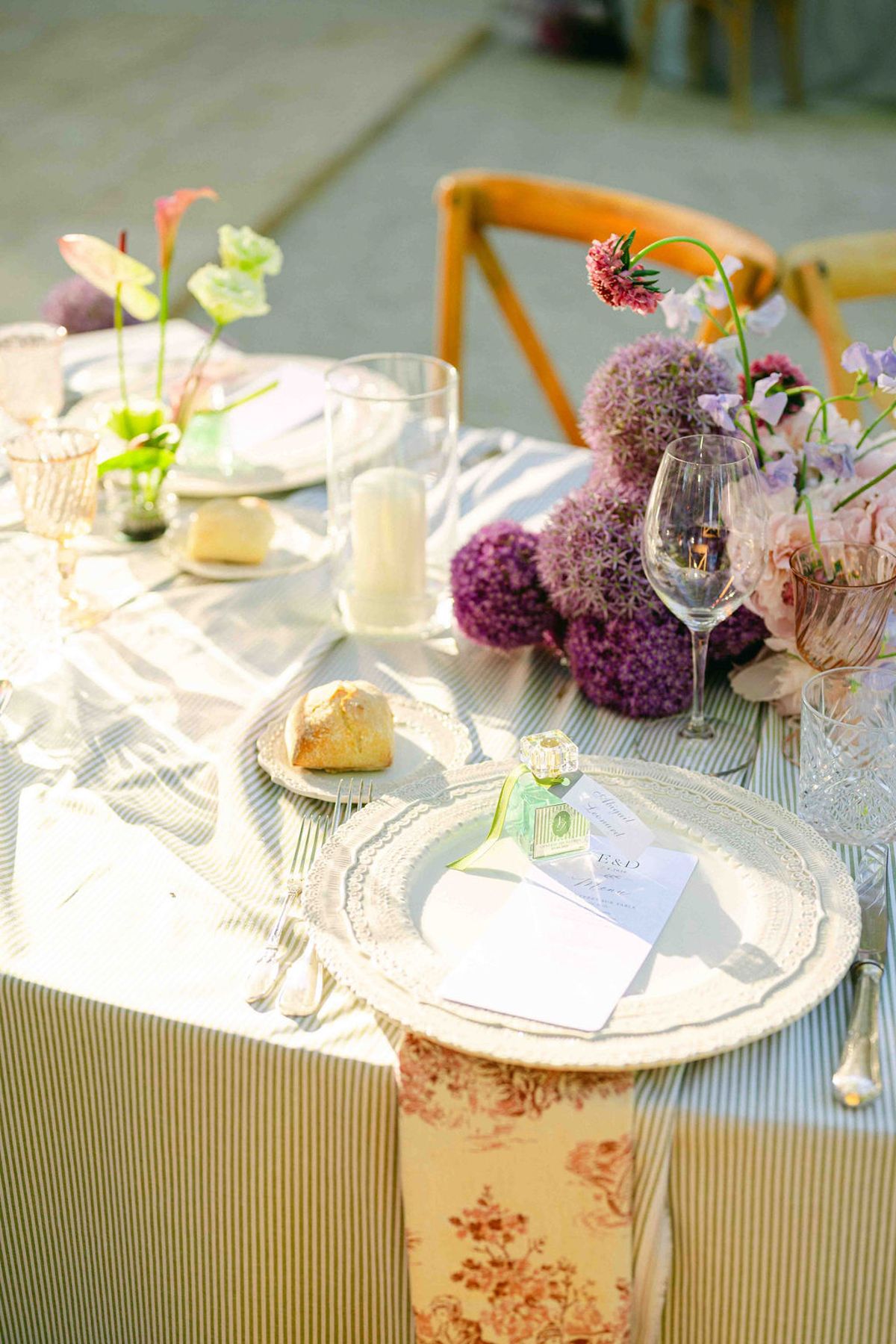 A dining table set with plates, glasses and a bread roll, with purple and pink flowers and a floral napkin.
