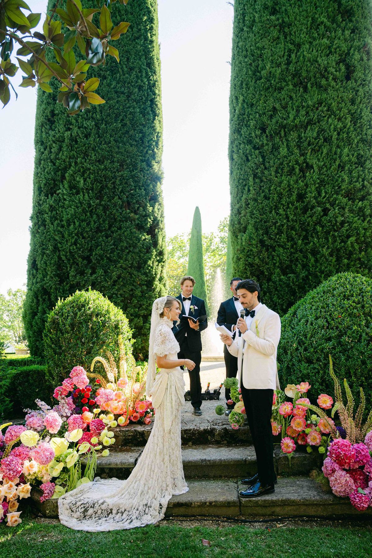 The bride and groom stand facing each other on garden steps with an officiant, surrounded by pink flowers and tall cypress trees.
