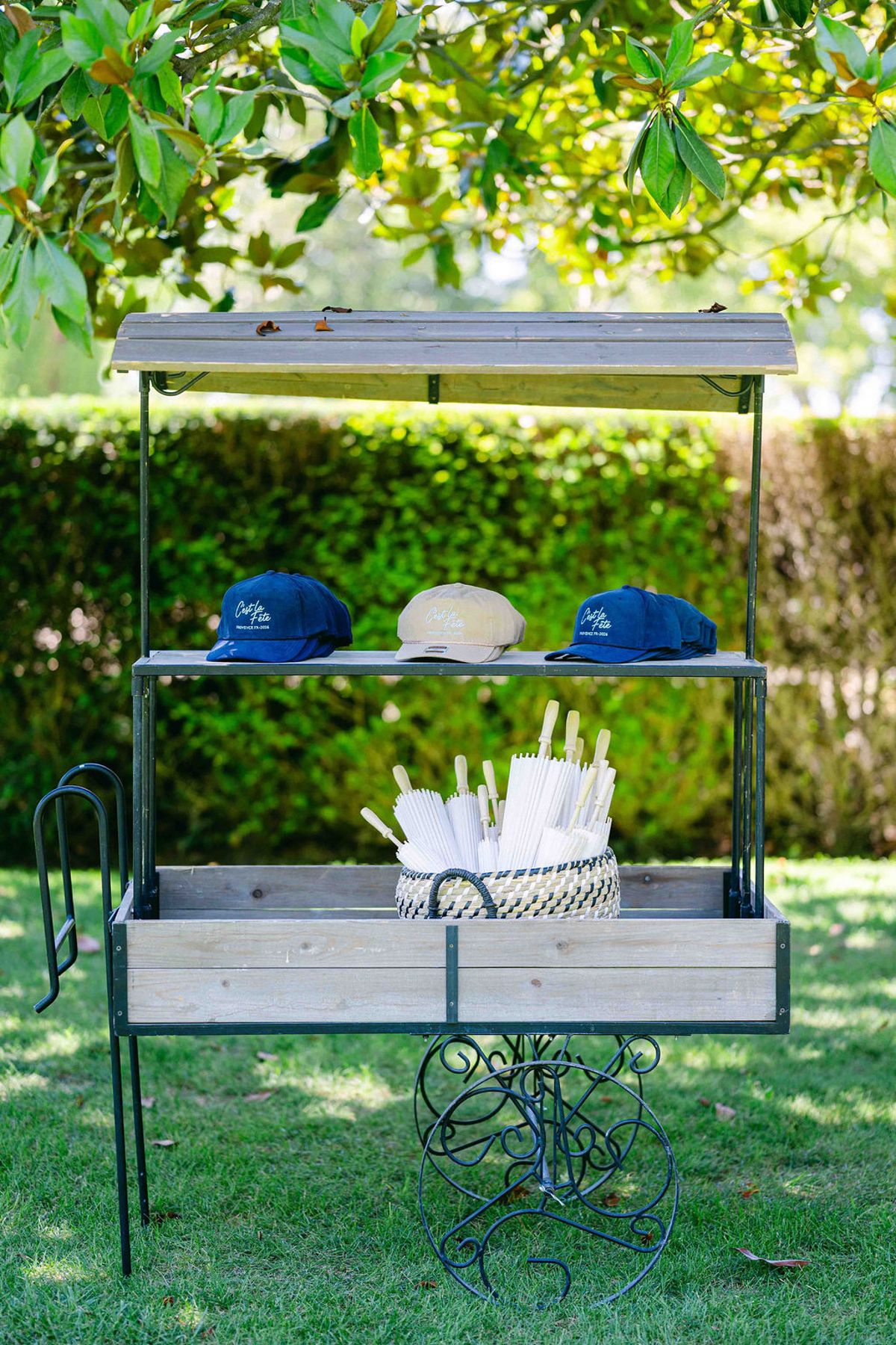 A small metal cart on a lawn displays blue and cream caps and rolled paper fans in front of a green hedge.