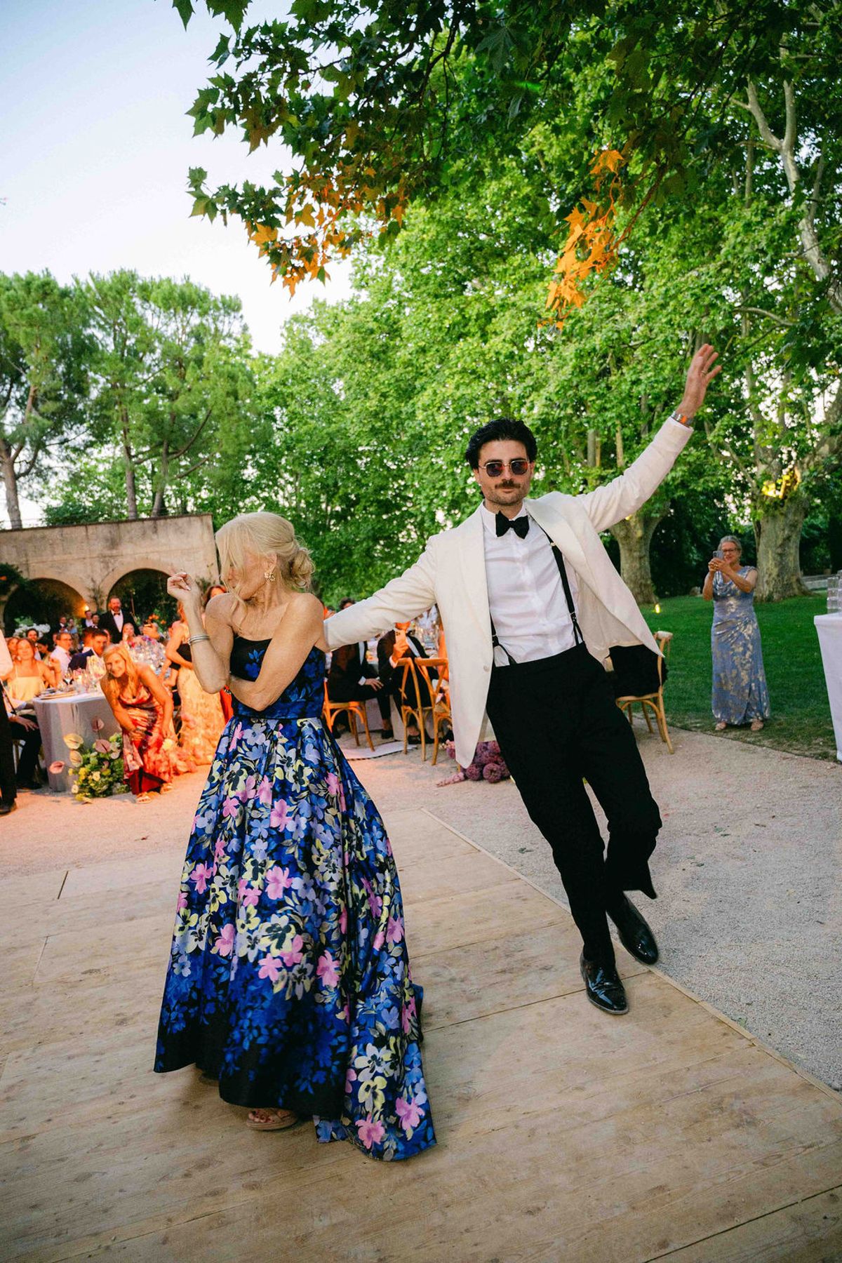 The groom in a white jacket and sunglasses dances with a woman in a blue floral gown on a terrace beneath trees.