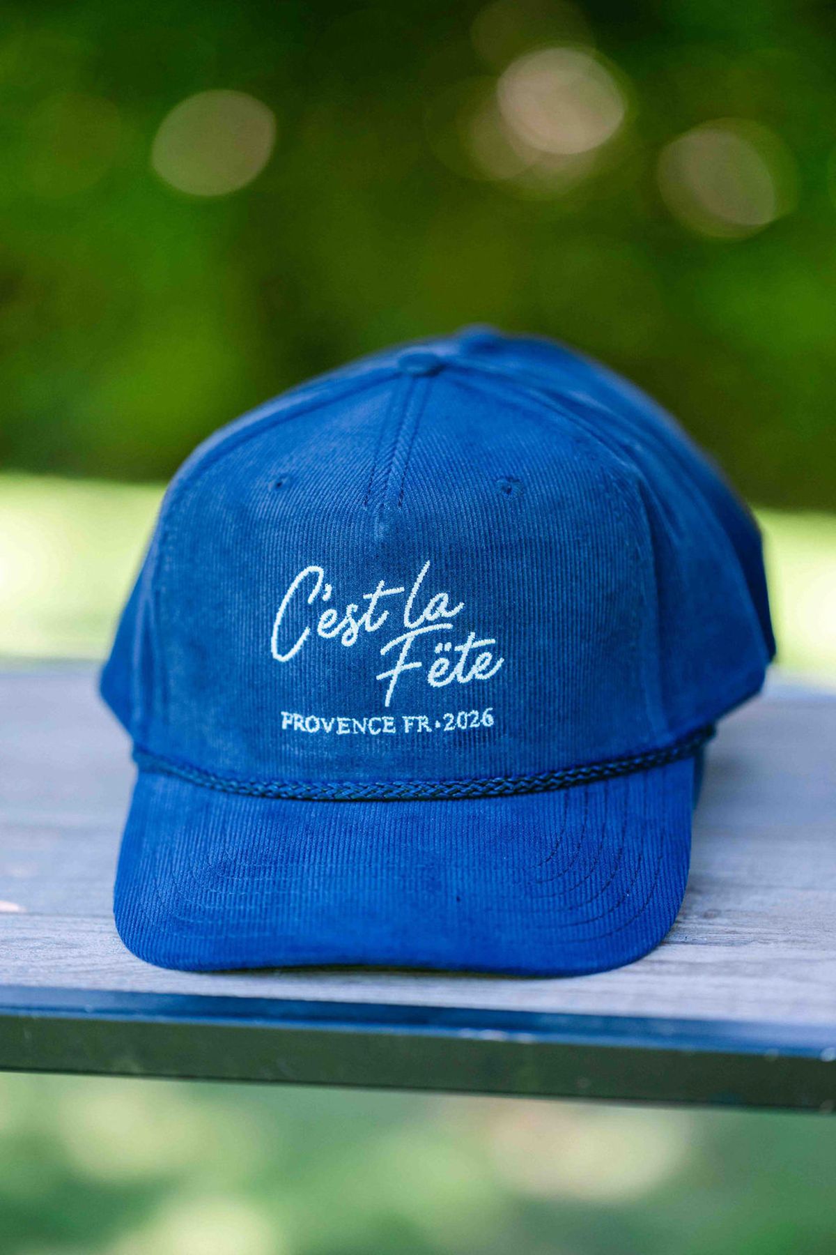 A blue corduroy cap with white script lettering rests on a wooden surface against a blurred green background.