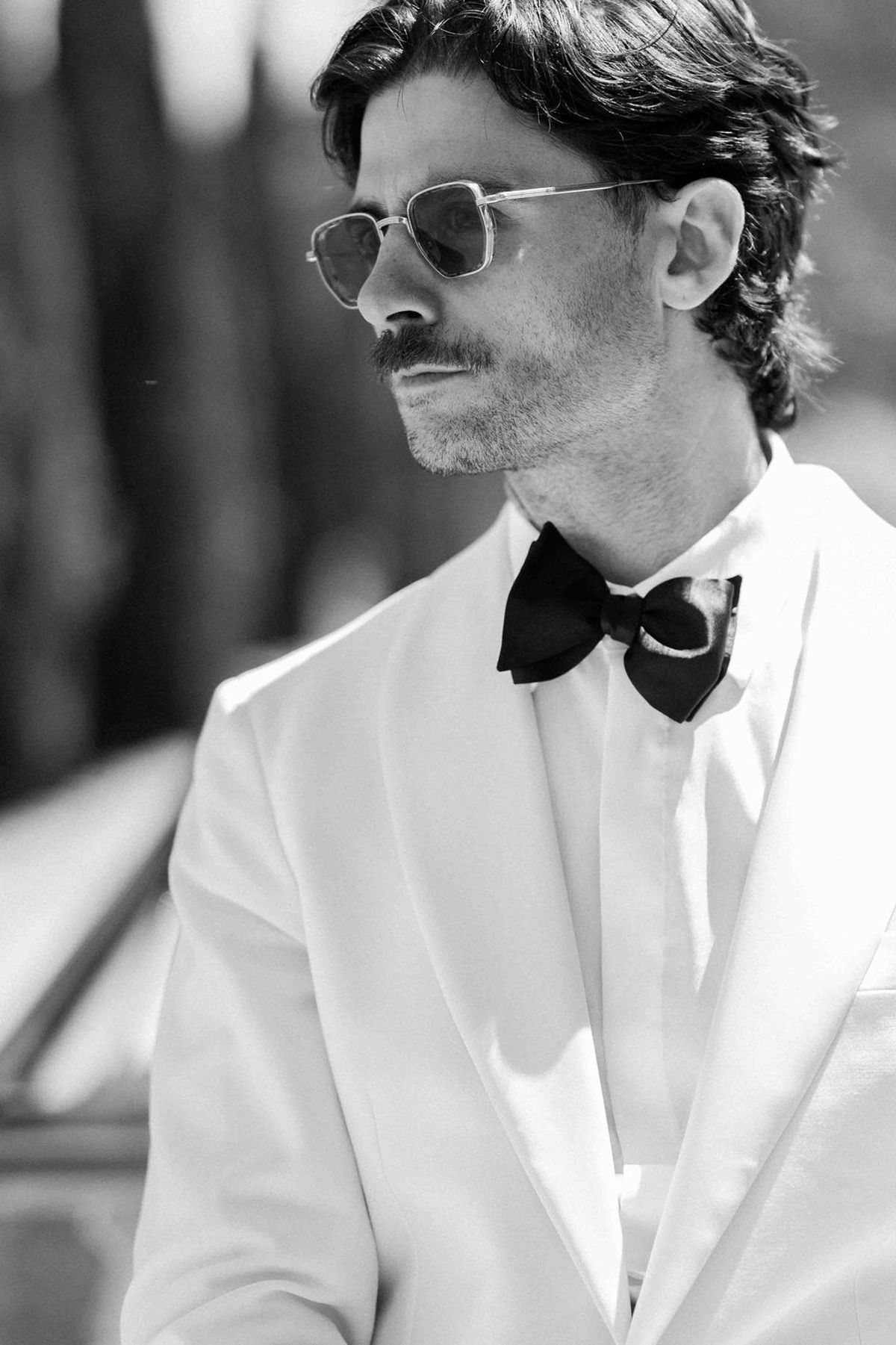 In black and white, the groom in round sunglasses, a white dinner jacket and black bow tie looks to one side.