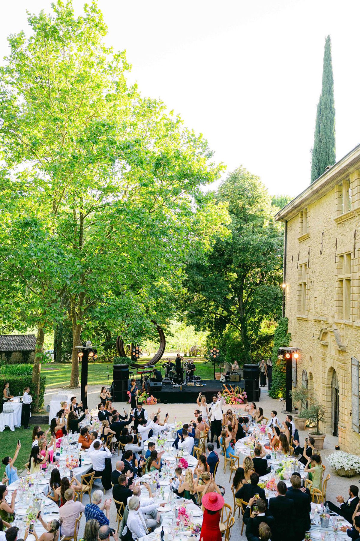 Guests sit at round tables in a sunlit courtyard beside a stone chateau, with trees overhead and a stage in the distance.