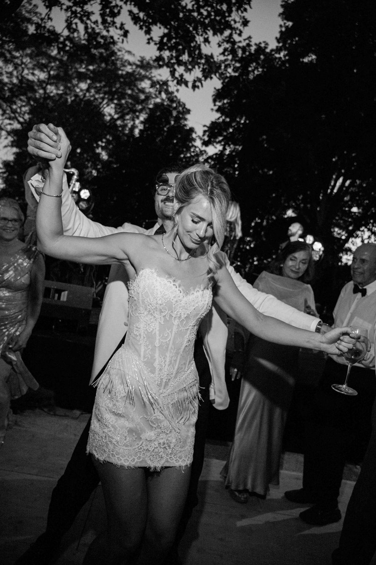 In black and white, a woman in a short lace dress dances with her arms raised, surrounded by guests.