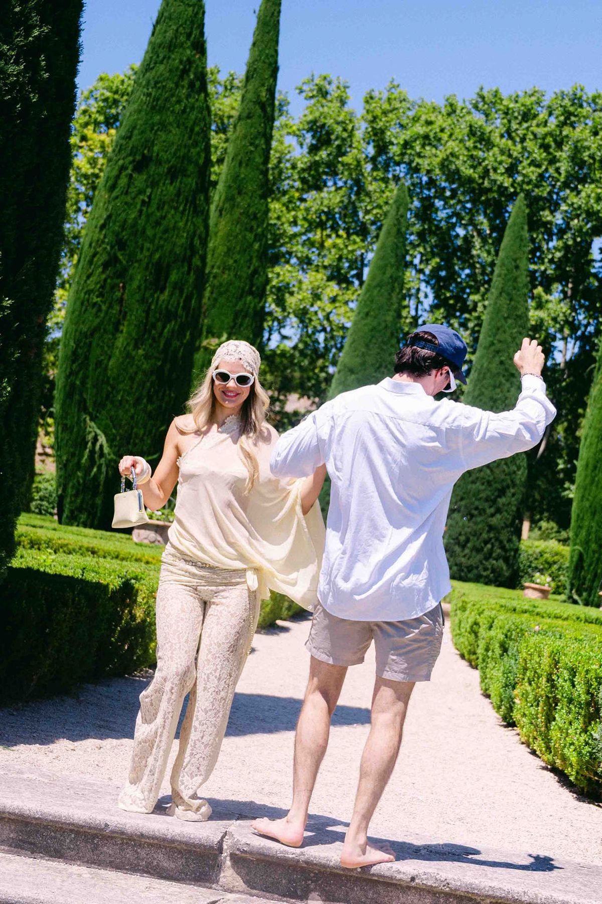 The couple walk playfully down stone steps in a garden of clipped hedges and tall cypress trees.