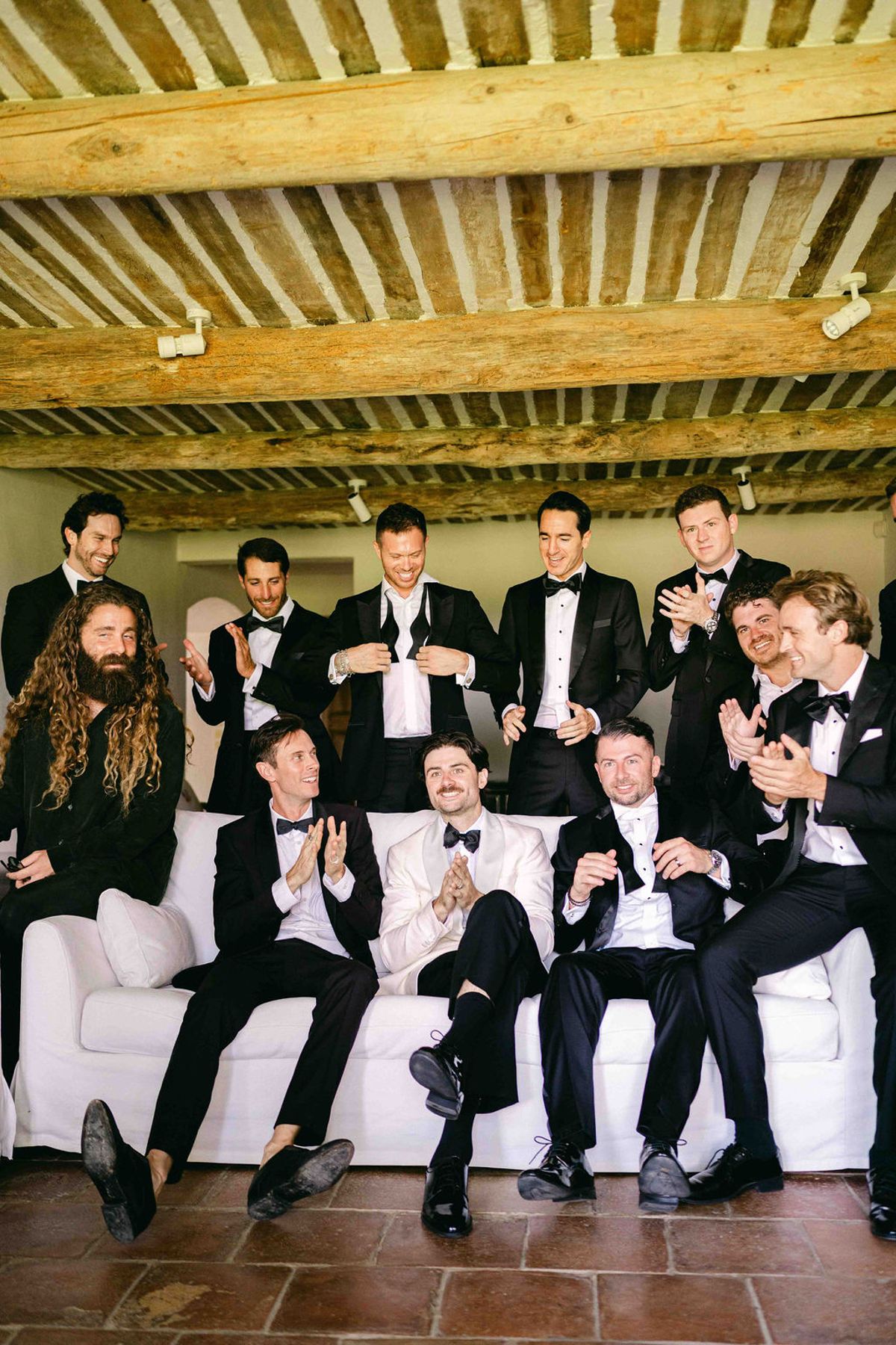 A large group of men in black tuxedos sit and stand around a white sofa, laughing in a room with a beamed ceiling.