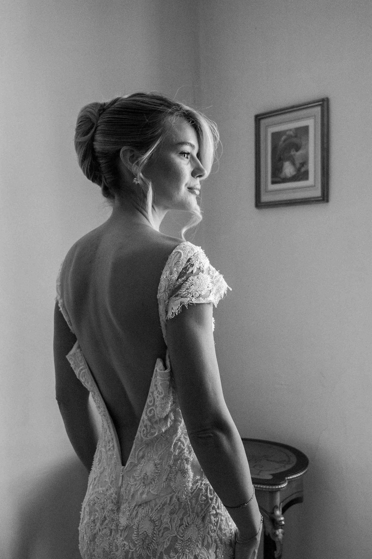 In black and white, a bride in a lace open-backed dress with her hair up looks to one side beside a window.